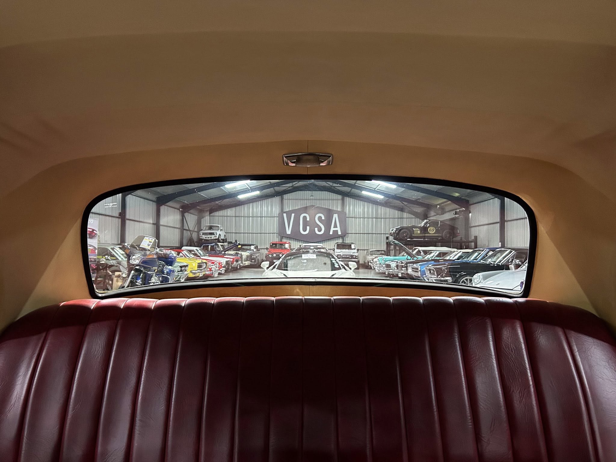 View from inside a vintage car showing a showroom full of classic cars, with a VCSA sign visible through the rear window.