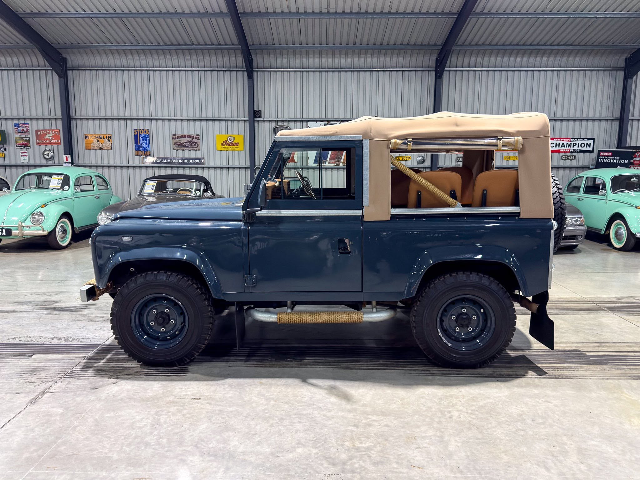 Inside a vintage car showroom with a dark blue off-road vehicle in the foreground and mint green classic cars along a corrugated metal wall lined with vintage posters
