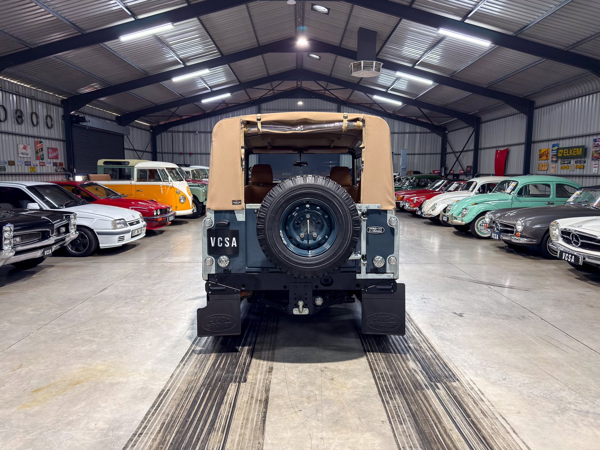 Rear view of a beige soft-top SUV with a mounted spare tire, inside a large vintage car showroom filled with colorful classic cars on both sides.