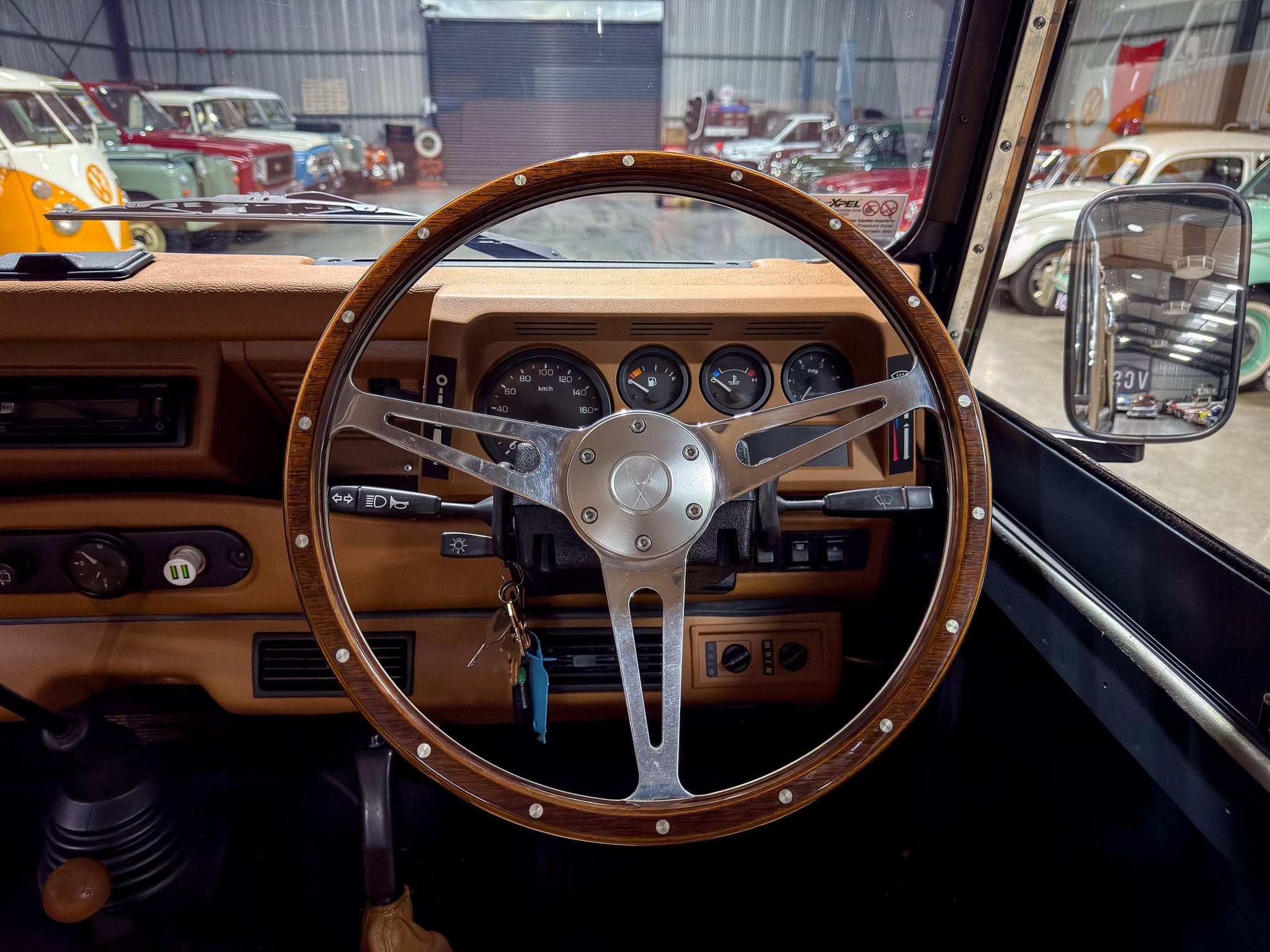Inside a vintage car cockpit with a wood-rimmed steering wheel and beige dashboard, showing classic gauges and switches while a garage full of vintage cars appears through the windshield.