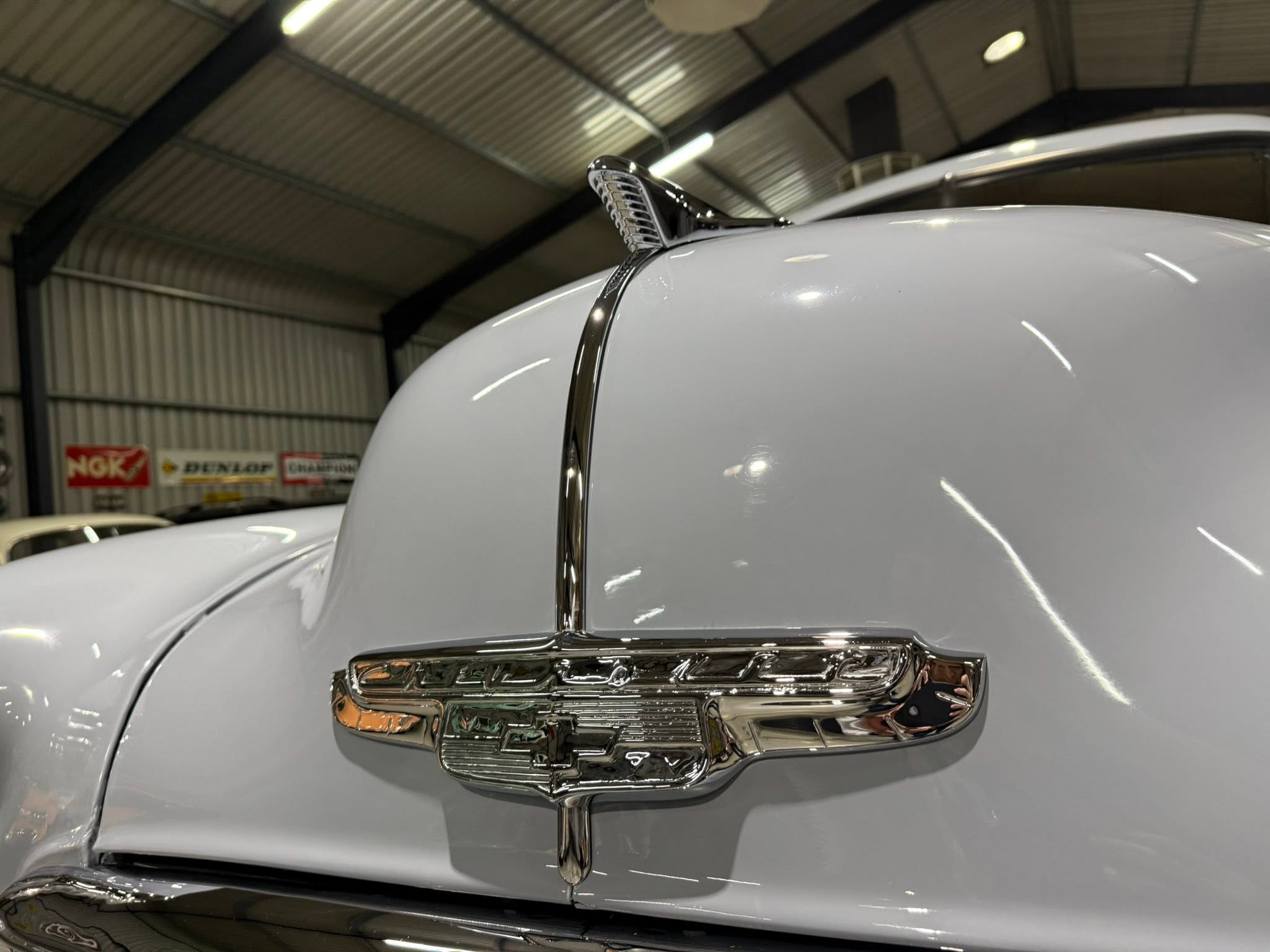 Close-up of a vintage car hood ornament and chrome grille on a light gray car indoors, a showroom backdrop behind it.