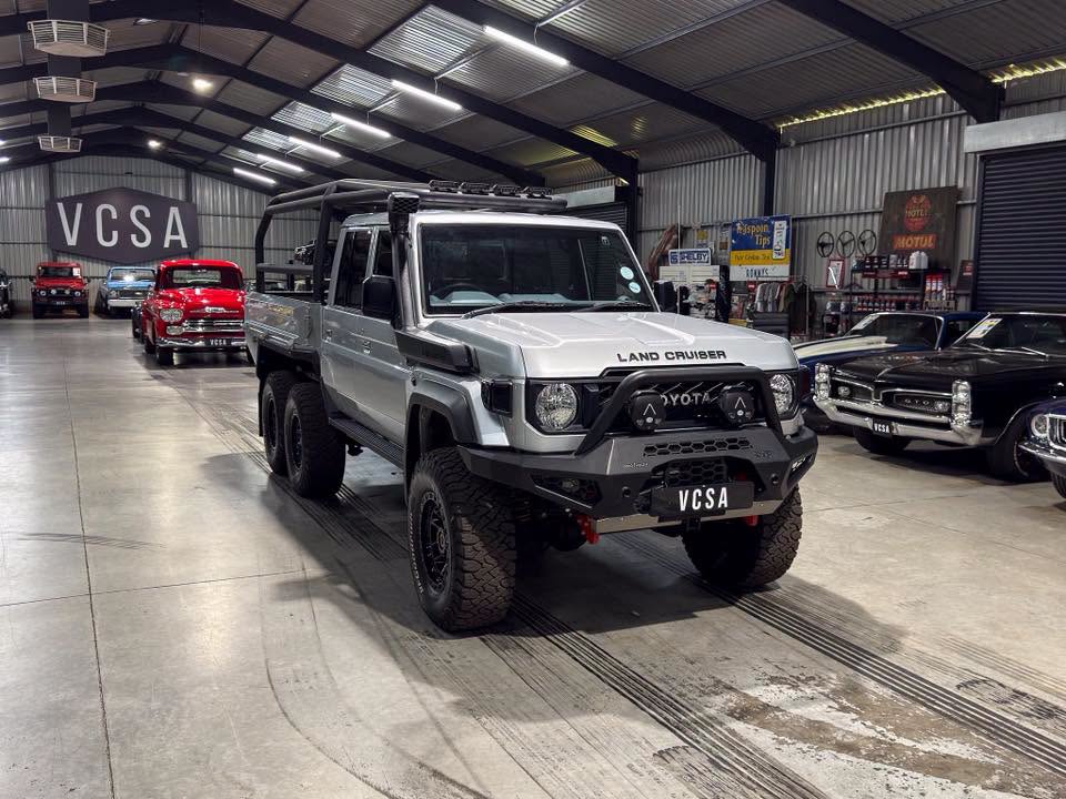 Gray Toyota Land Cruiser with a heavy bull bar and lifted tires in a well-lit showroom, classic cars visible in the background (VCSA signs).