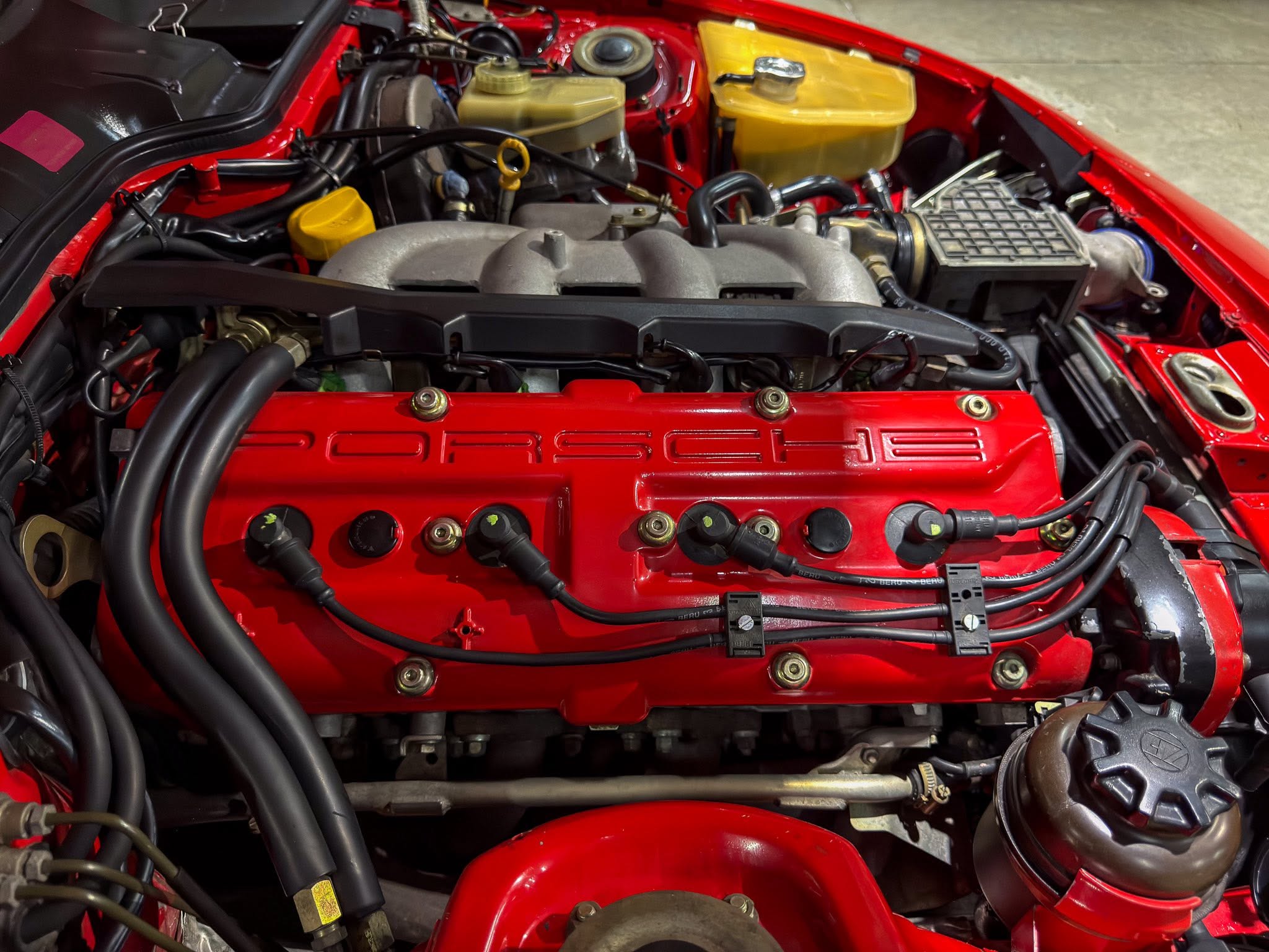 Red Porsche engine bay with exposed intake manifold, black hoses, and yellow coolant reservoir visible in the back left.