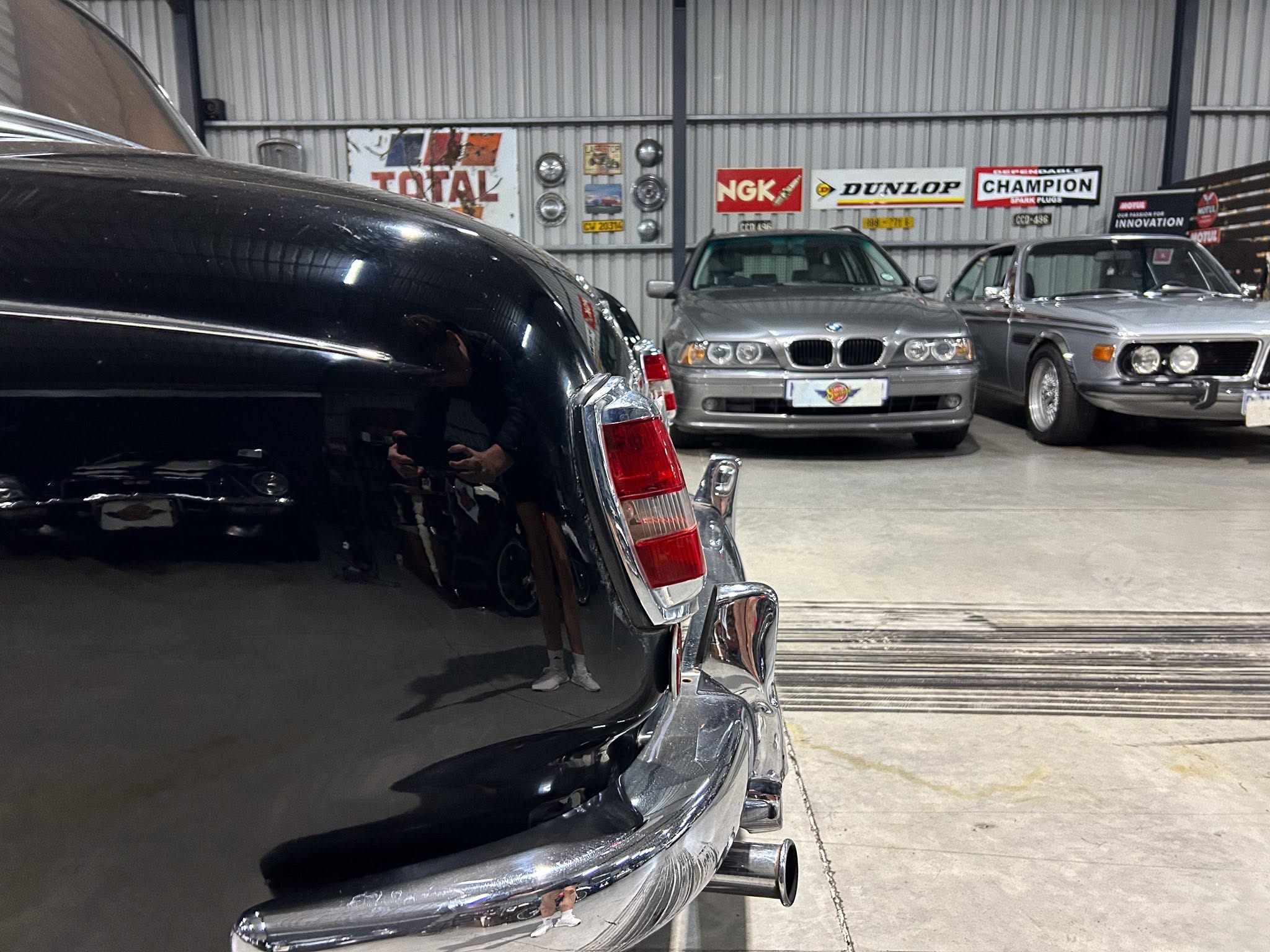 Close-up of a glossy black car bumper in a showroom-like garage, with two silver classic cars and vintage signs in the background.