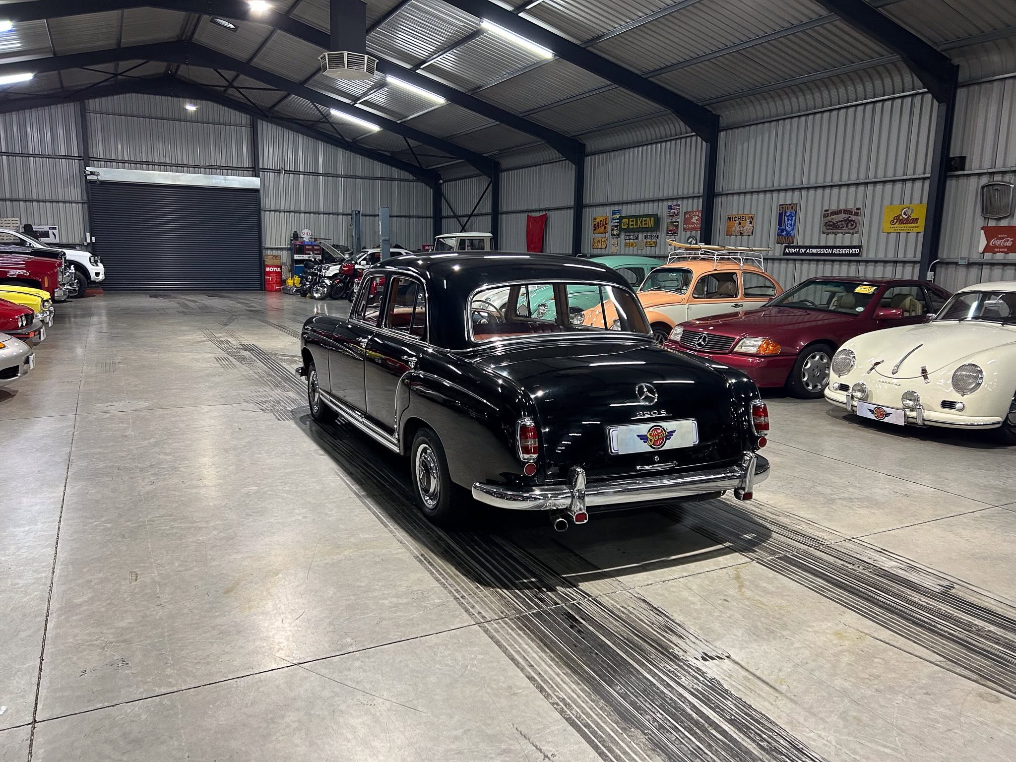 Indoor showroom of vintage cars lined up along metal walls and banners in a large warehouse.