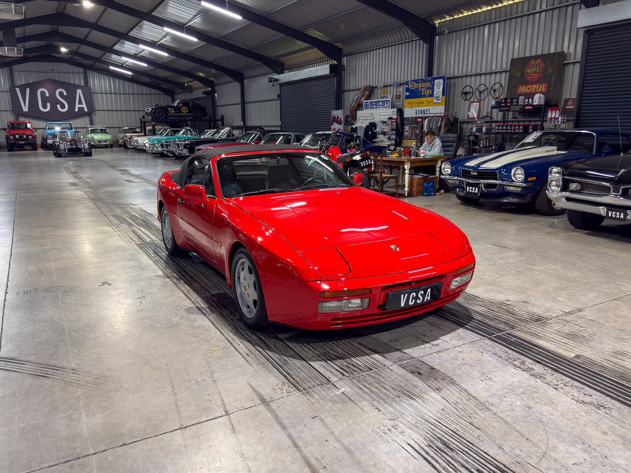 Interior of a vintage car workshop with a bright red convertible in the foreground and a row of classic cars along the left wall; a large VCSA sign on the back wall.