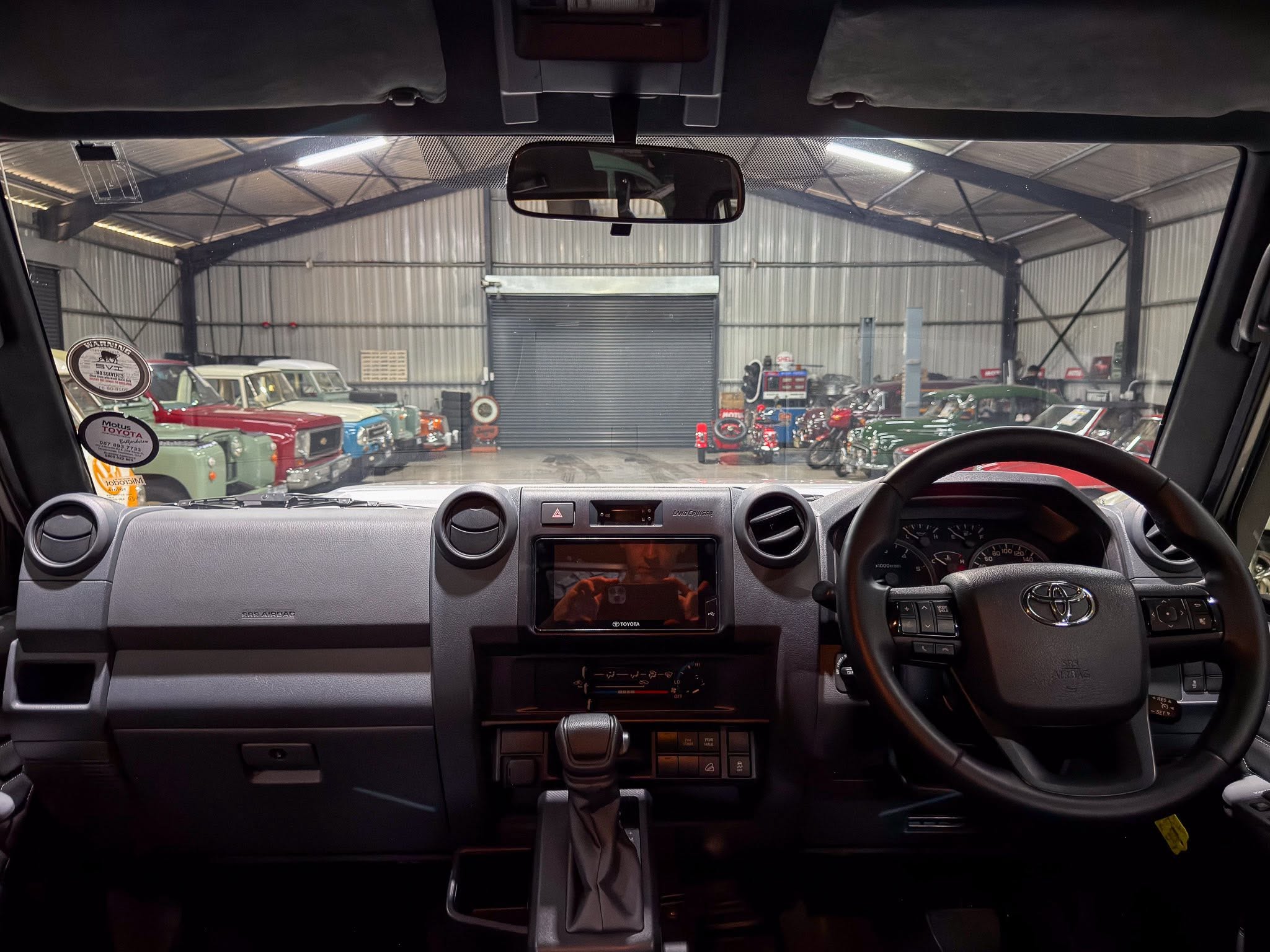 View from the driver seat of a modern car with steering wheel and center console, looking out at a warehouse of vintage cars through the windshield.
