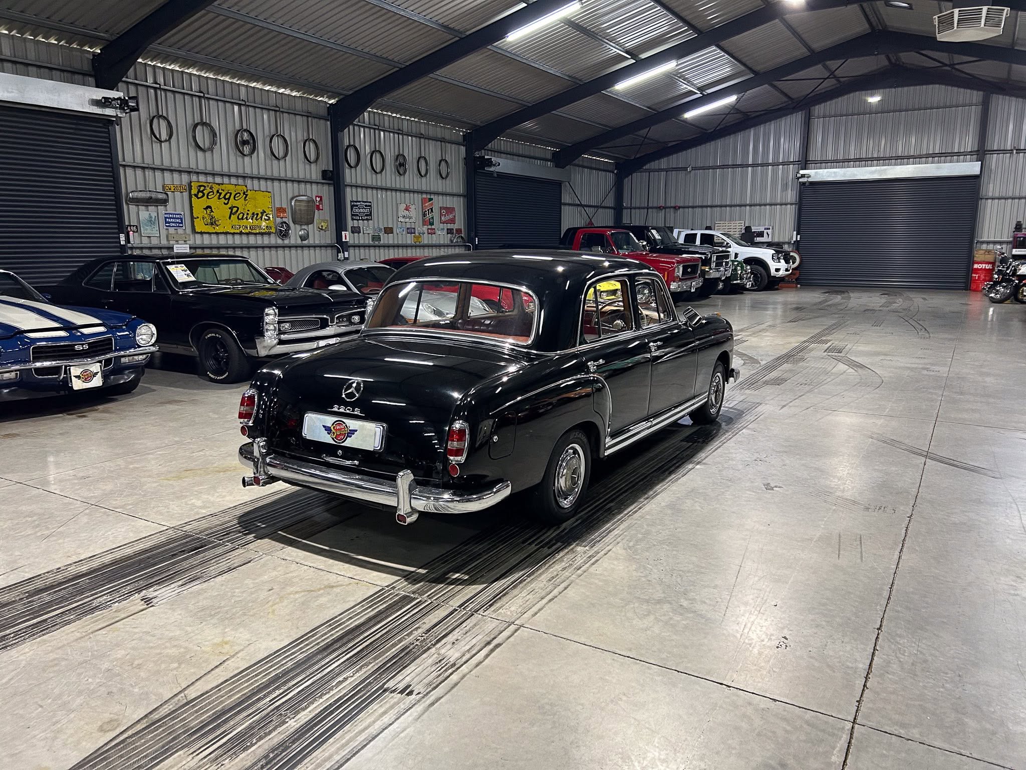 Indoor metal-clad garage showroom with several classic cars, including a black Mercedes at the front and other vintage vehicles lined along the walls.
