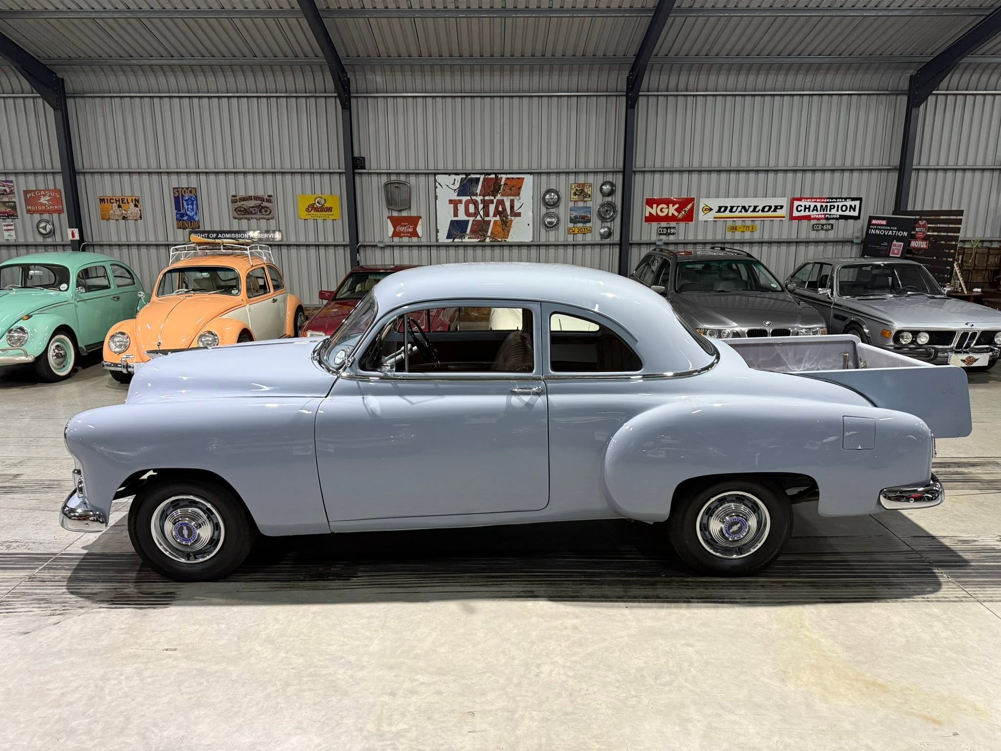 Showroom scene with a light blue vintage car in the foreground and pastel-colored classic cars against a wall of retro signs and posters.