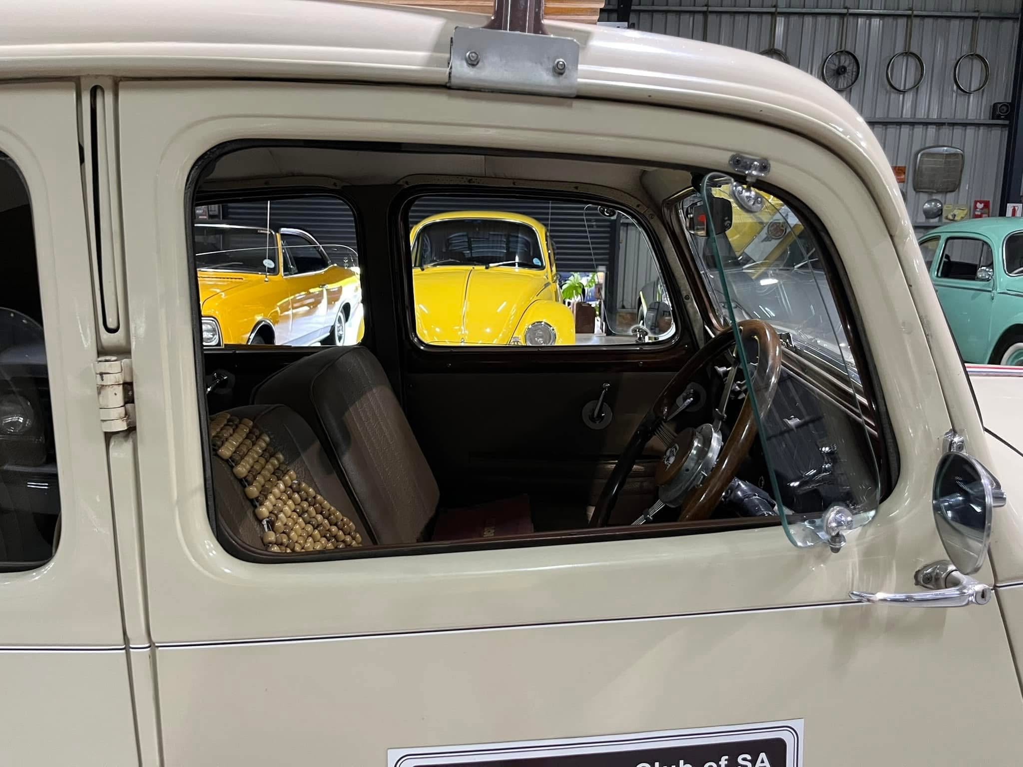 Inside a vintage car with beige upholstery and a beaded seat cover, seen through the open window at a showroom with yellow classic cars outside.