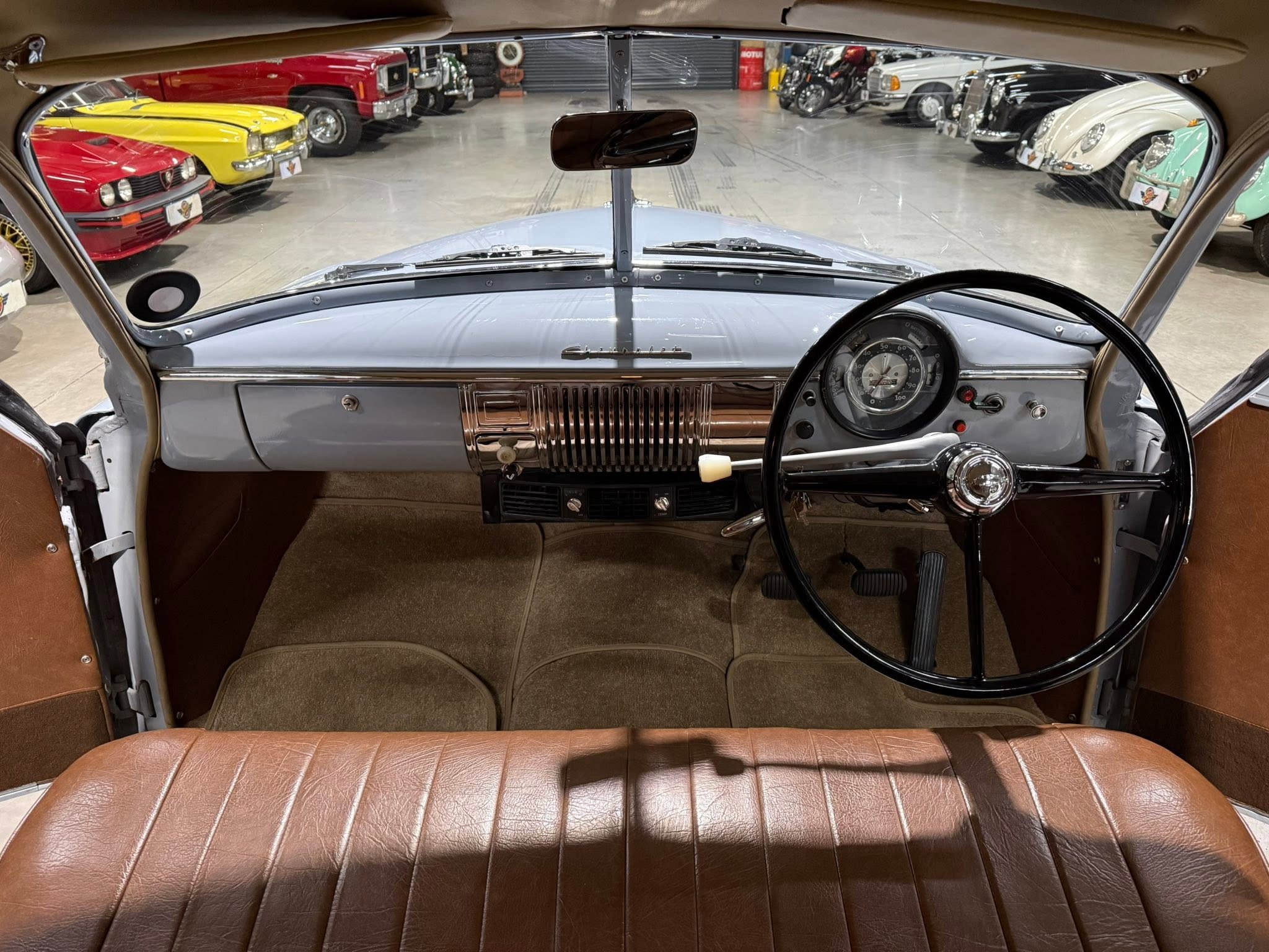 View from the driver’s seat inside a vintage car, looking out at a showroom filled with colorful classic cars parked in two rows behind a gray dashboard and brown upholstery.