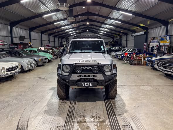 White Toyota Land Cruiser centered in a large indoor showroom, flanked by classic cars on both sides.