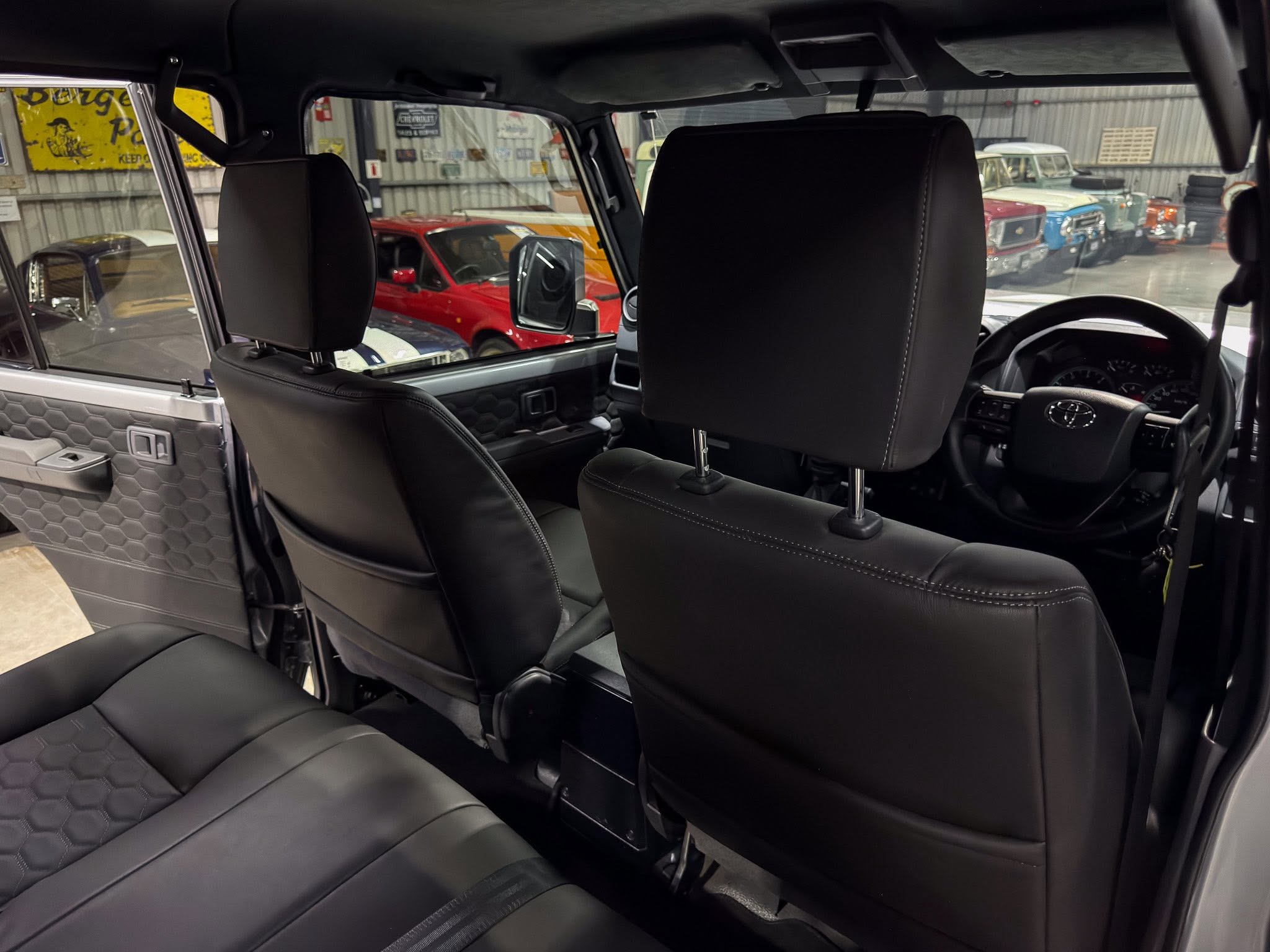 Backseat interior of a black leather SUV with visible headrests and a view of the driver’s seat, steering wheel, and dashboard; large glass showroom windows show vintage cars outside.