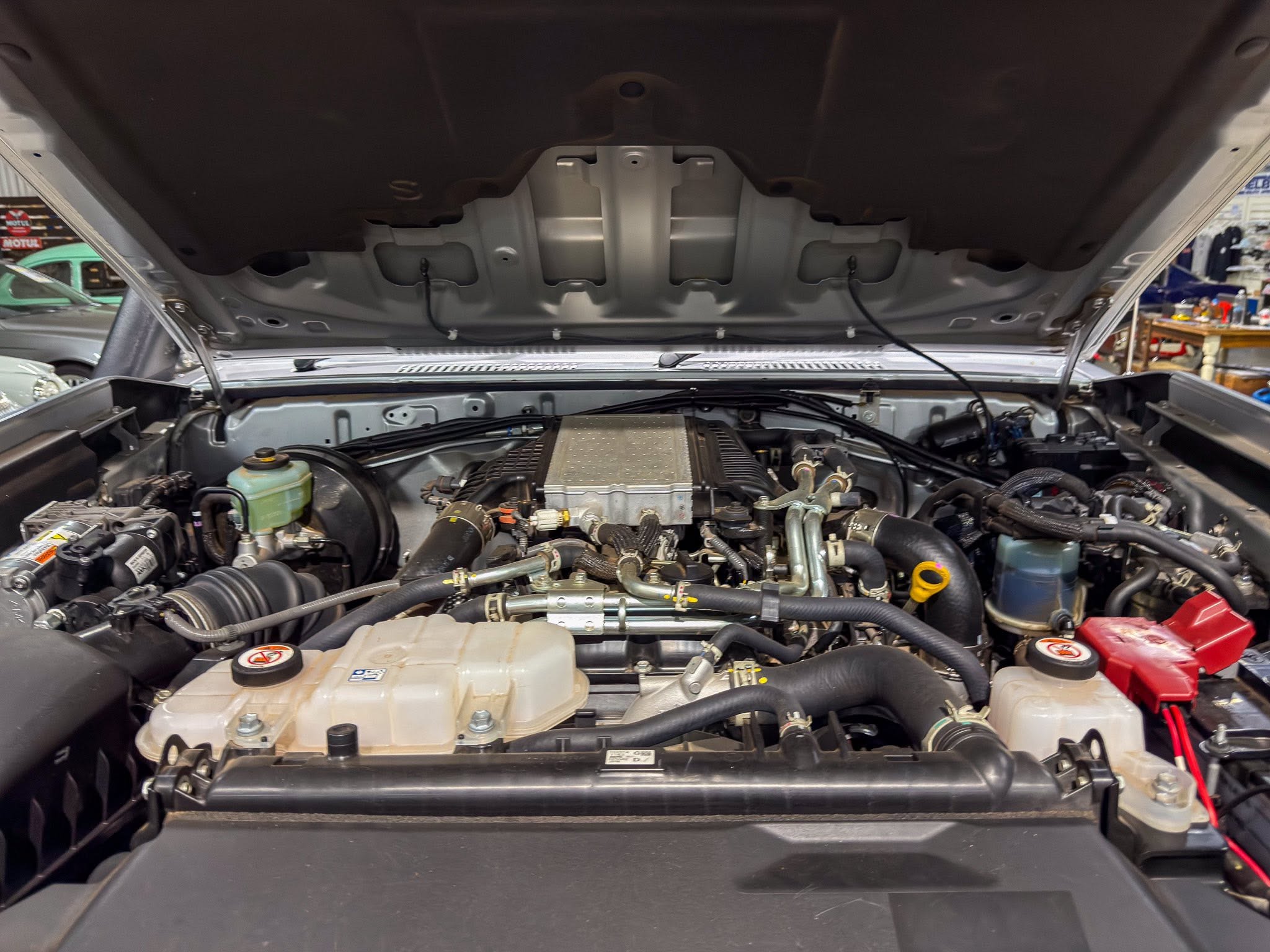 Under-hood view of a vehicle with the hood raised, showing the engine, hoses, and fluid reservoirs in a workshop.