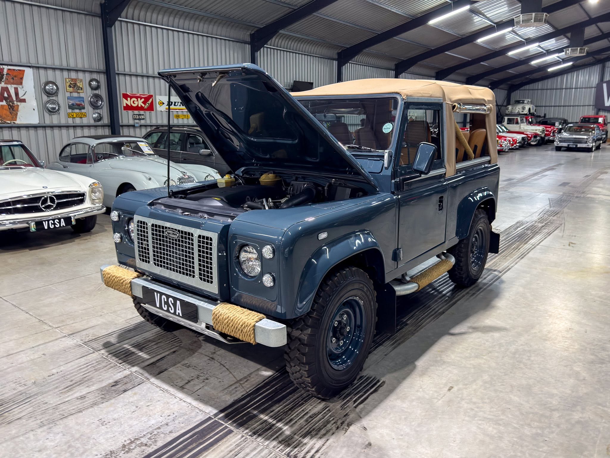 Blue classic Land Rover Defender with the hood up in a vintage car showroom, surrounded by other classic cars inside a metal-walled venue.