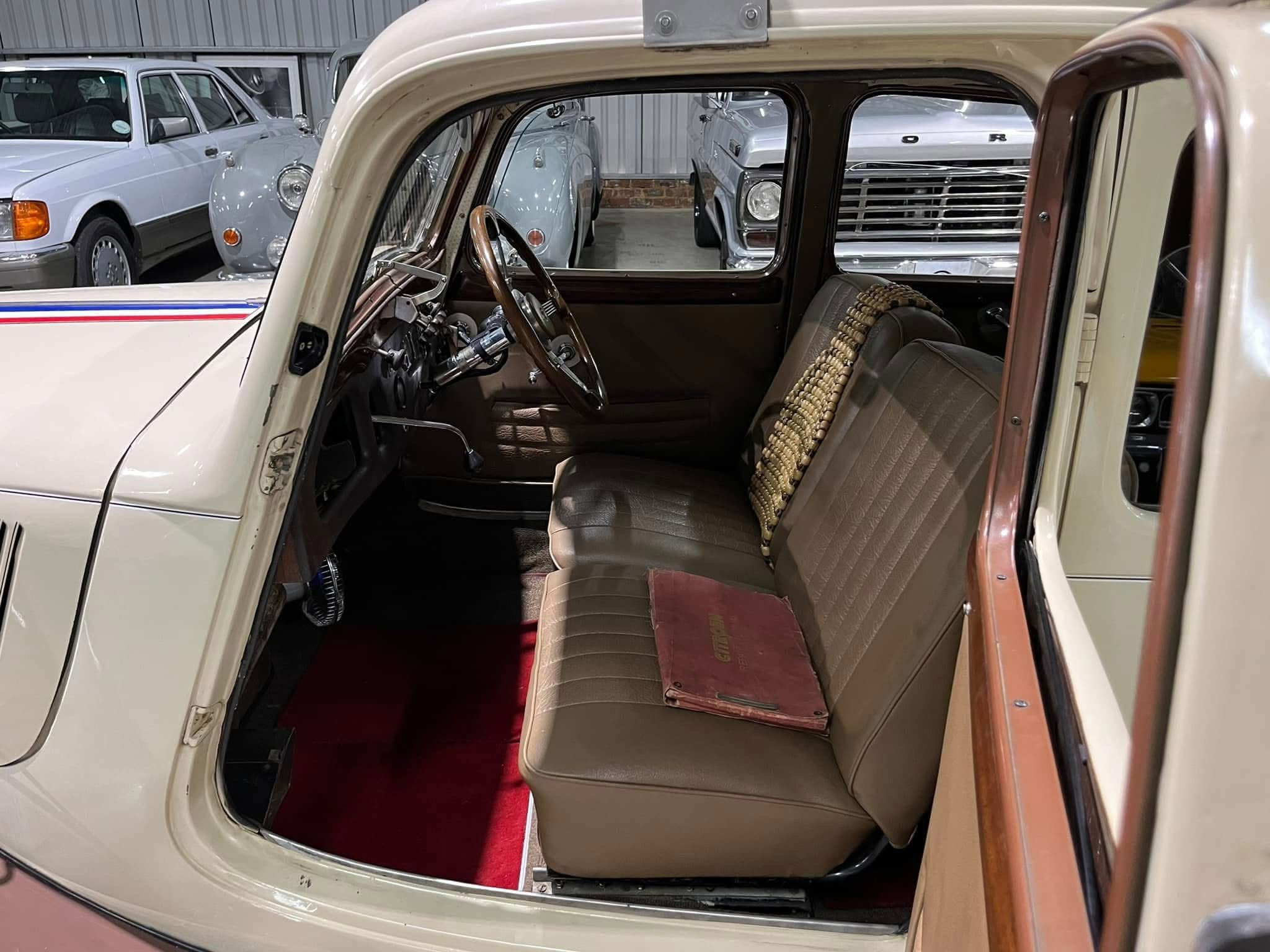 Open-door view of a vintage beige car interior with a wooden steering wheel and tan seats; red carpet on the floor.