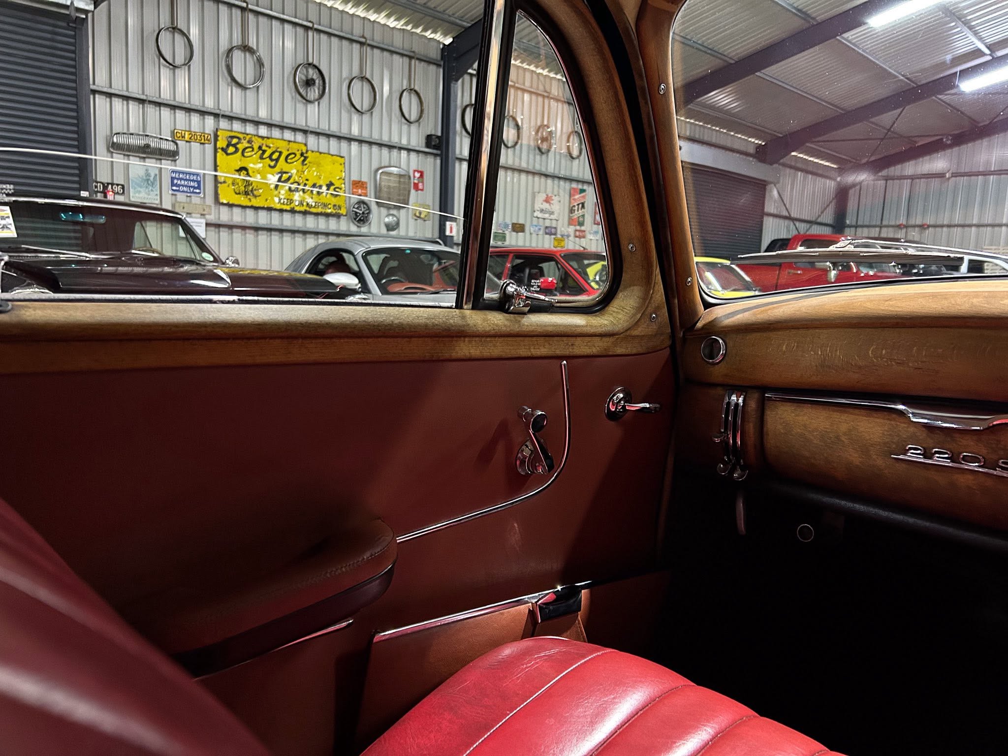 Interior of a vintage car with red leather seats and wood trim, looking out the side window at a workshop with classic cars and retro signage on the wall