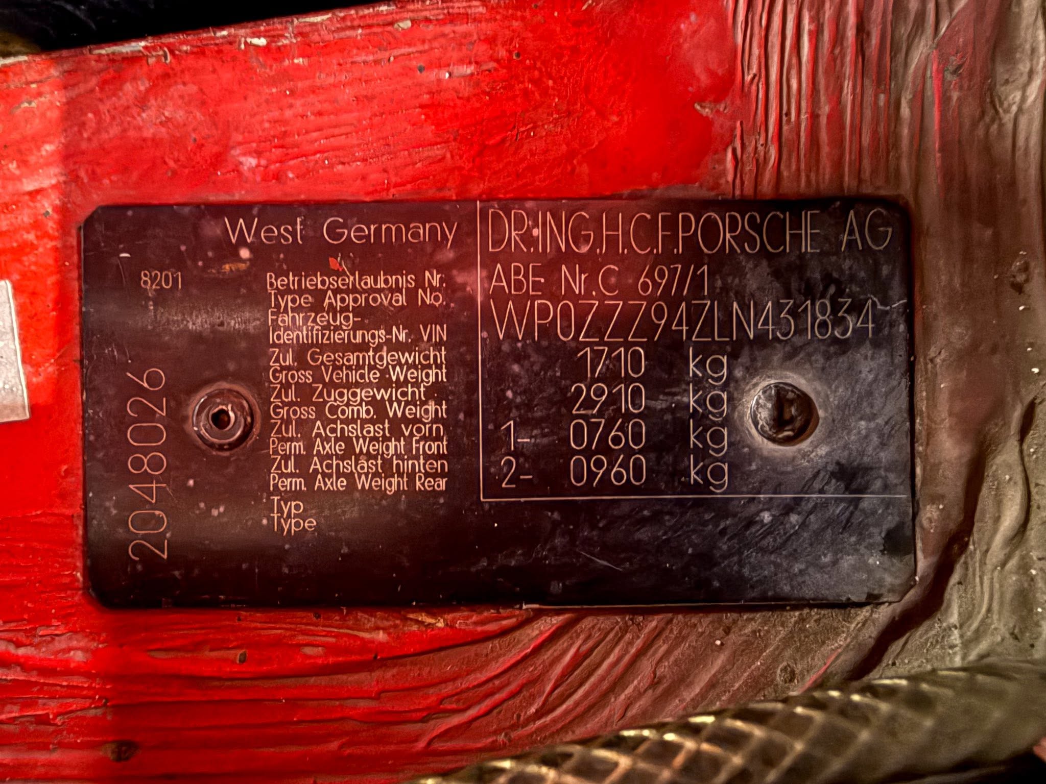 Metal vehicle identification plate on a red painted surface, showing West Germany, Porsche AG, and weight/ VIN details.