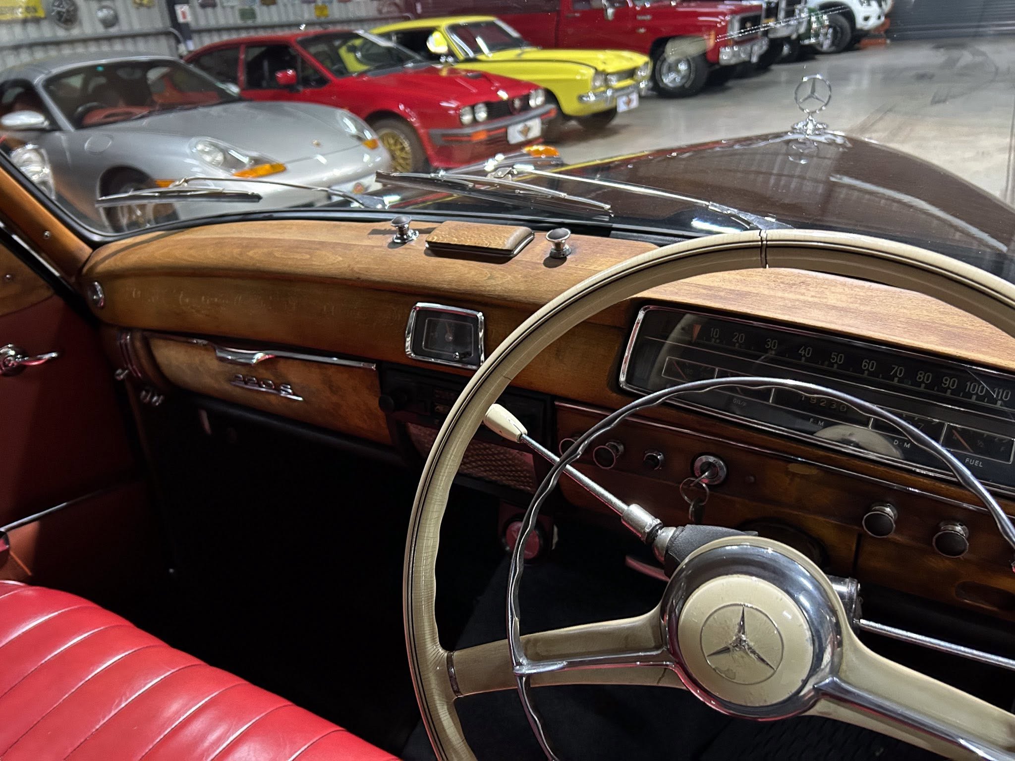 View of a classic car interior with a wood dashboard, leather red seat, and chrome steering wheel centerpiece; showroom of vintage cars outside the windshield. Viewed from the driver’s seat, showing a wood-grain dash, analog gauges, and a Mercedes emblem on the steering wheel, with a lineup of classic cars in the background.