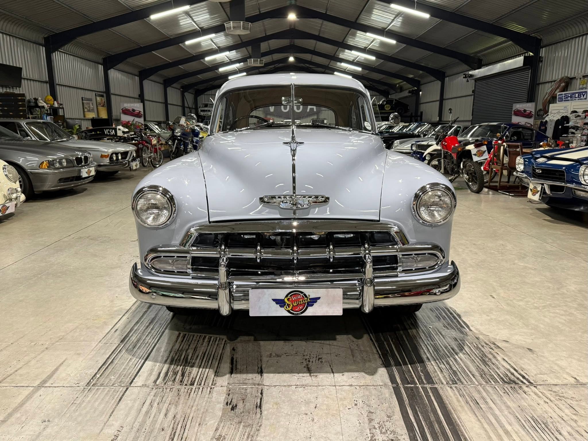 Front and center of a light blue vintage car inside a showroom filled with classic cars and motorcycles.