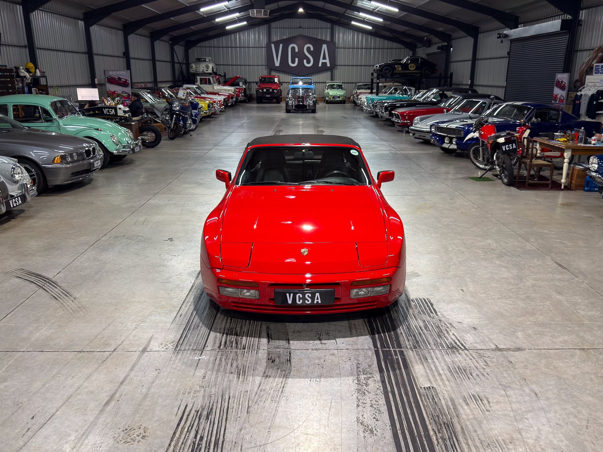 Front-on view of a red sports car in a large vintage car showroom, flanked by rows of classic cars and a VCSA sign in the back.