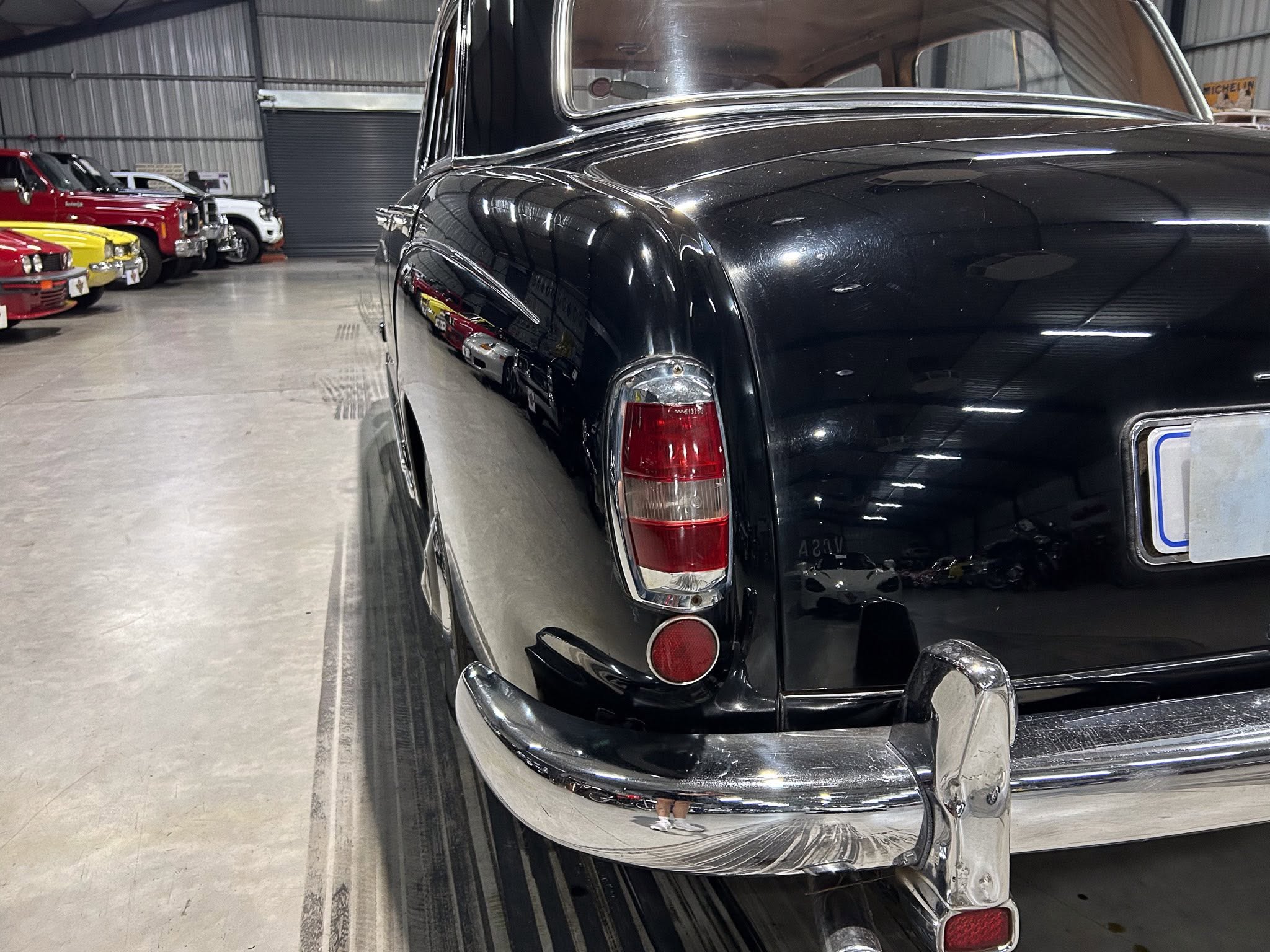 Close-up of a glossy black vintage car's rear quarter panel with chrome bumper in a showroom, other colorful cars in the background.