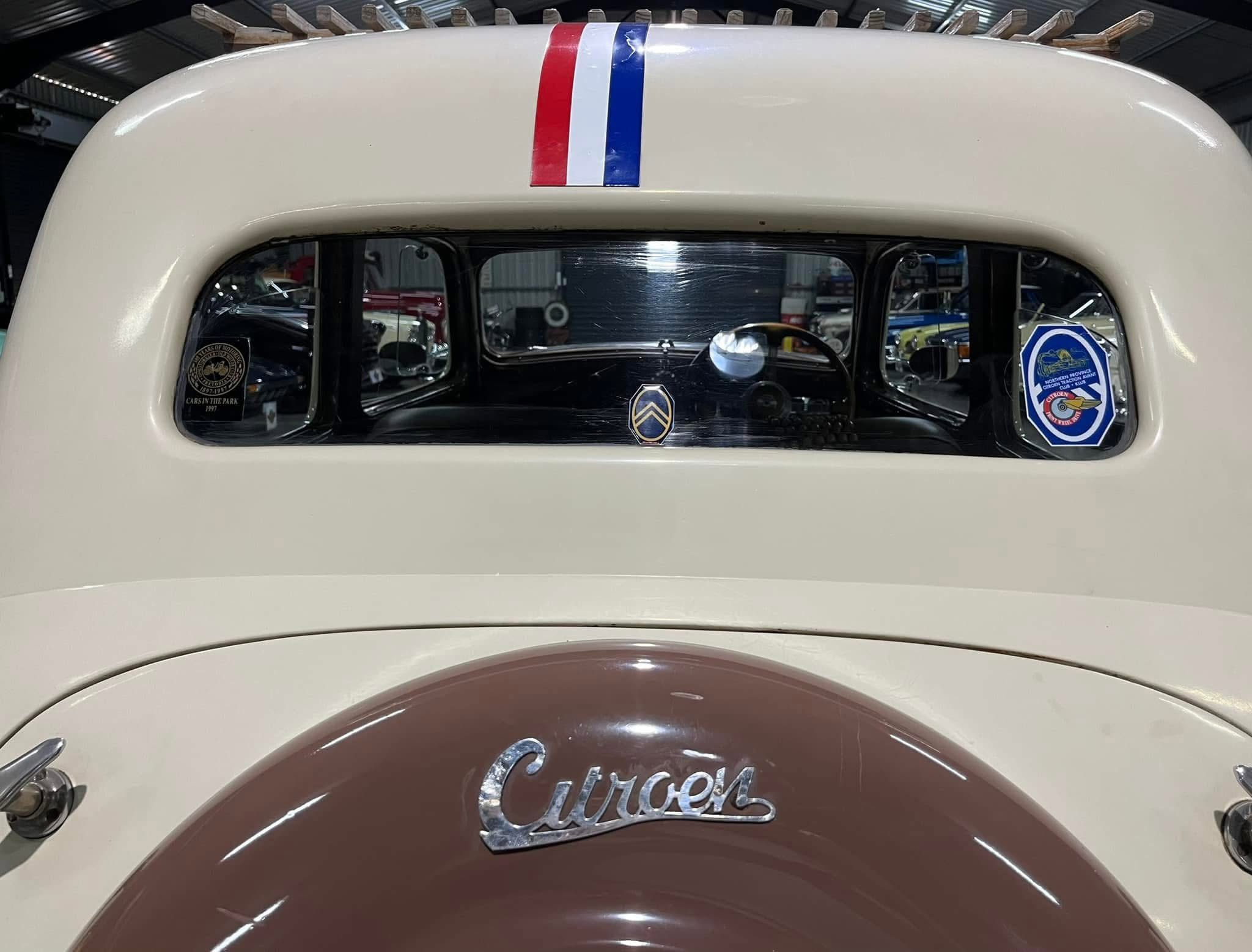 Rear view of a cream-colored vintage car with a red‑white‑blue stripe on the roof, visible through the rear window. Stickers and a Citroën emblem appear on the glass.
