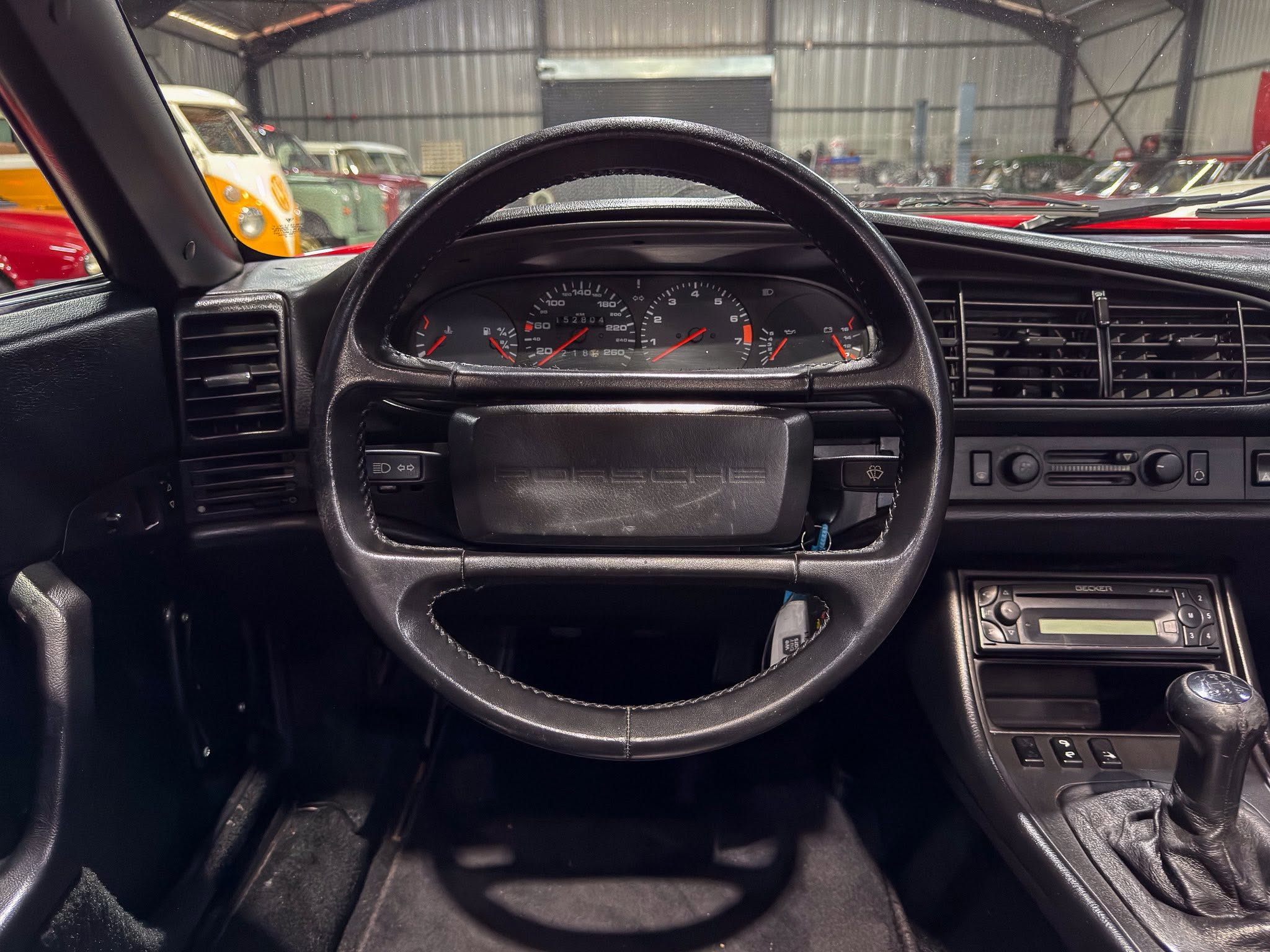 View from the driver's seat of a car interior, showing the steering wheel, instrument cluster, and center console in a garage full of classic cars outside the windshield.