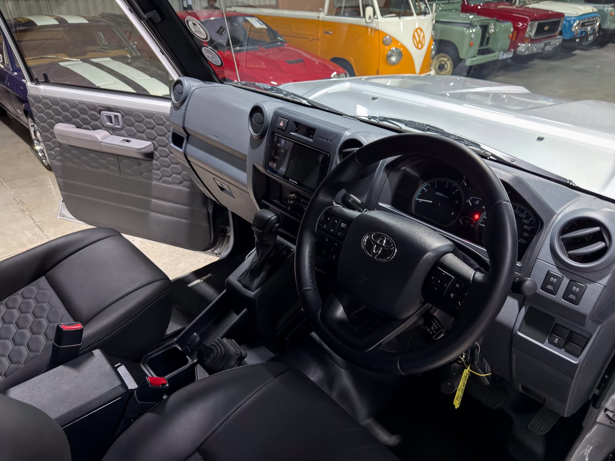 Front interior of a dark gray car showing the honeycomb-pattern door panel, steering wheel, center console, and a row of vintage vans outside the windshield.