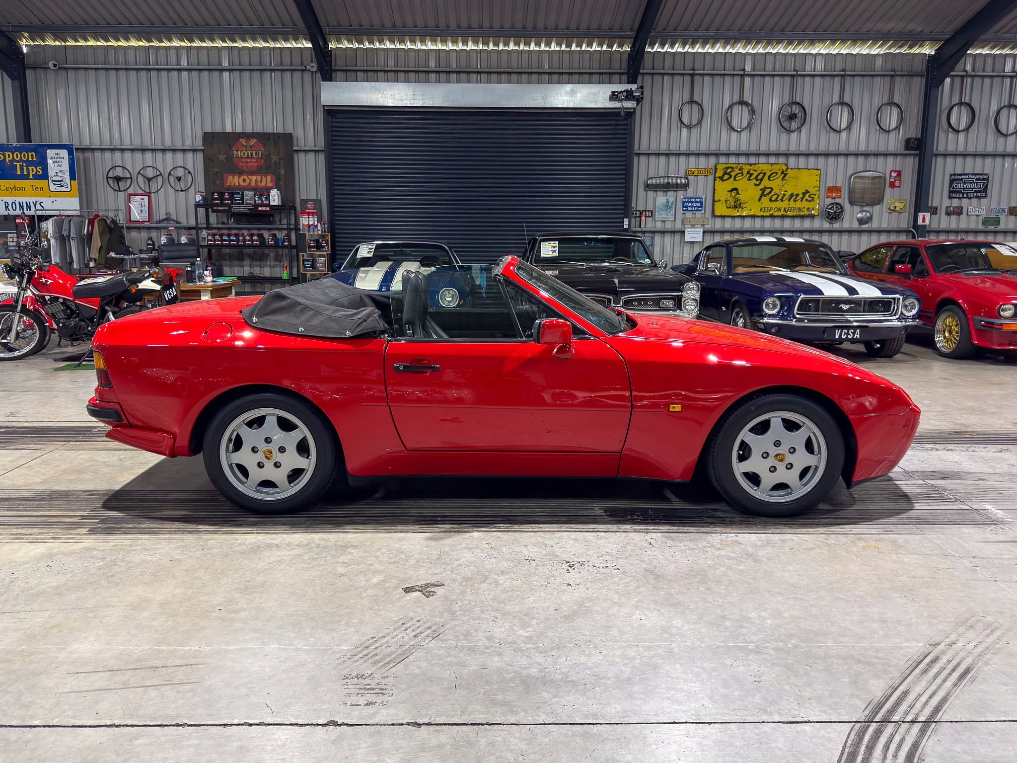 Red convertible with the top down parked inside a vintage car showroom, surrounded by classic cars and retro signs on metal walls.