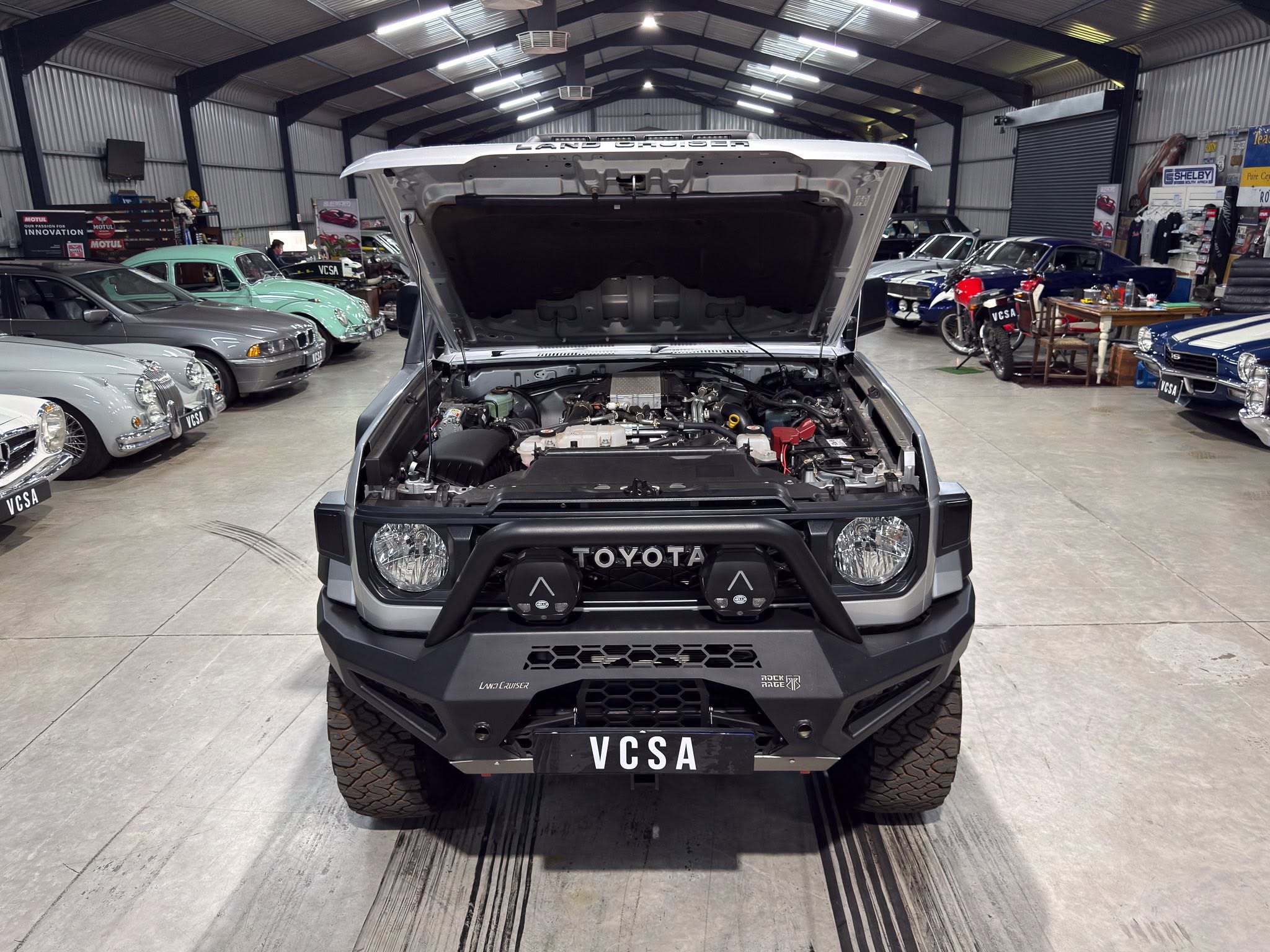 Front view of a rugged Toyota with hood open in a classic car showroom, surrounded by vintage cars and tires stacked along the walls.