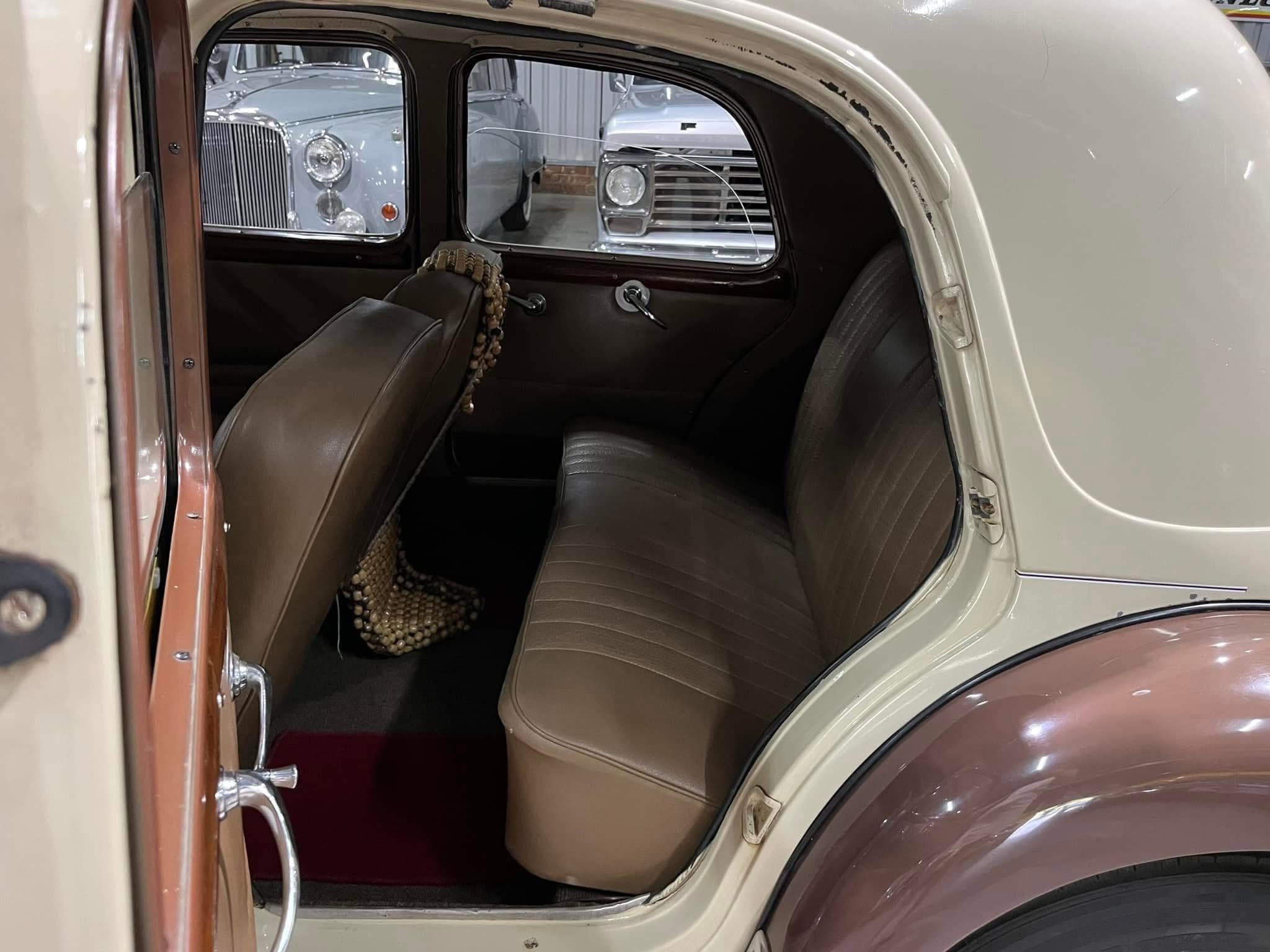 Interior of a vintage car with brown leather seats, a beaded seat cover, and chrome window frames; another classic car is visible through the side window outside.