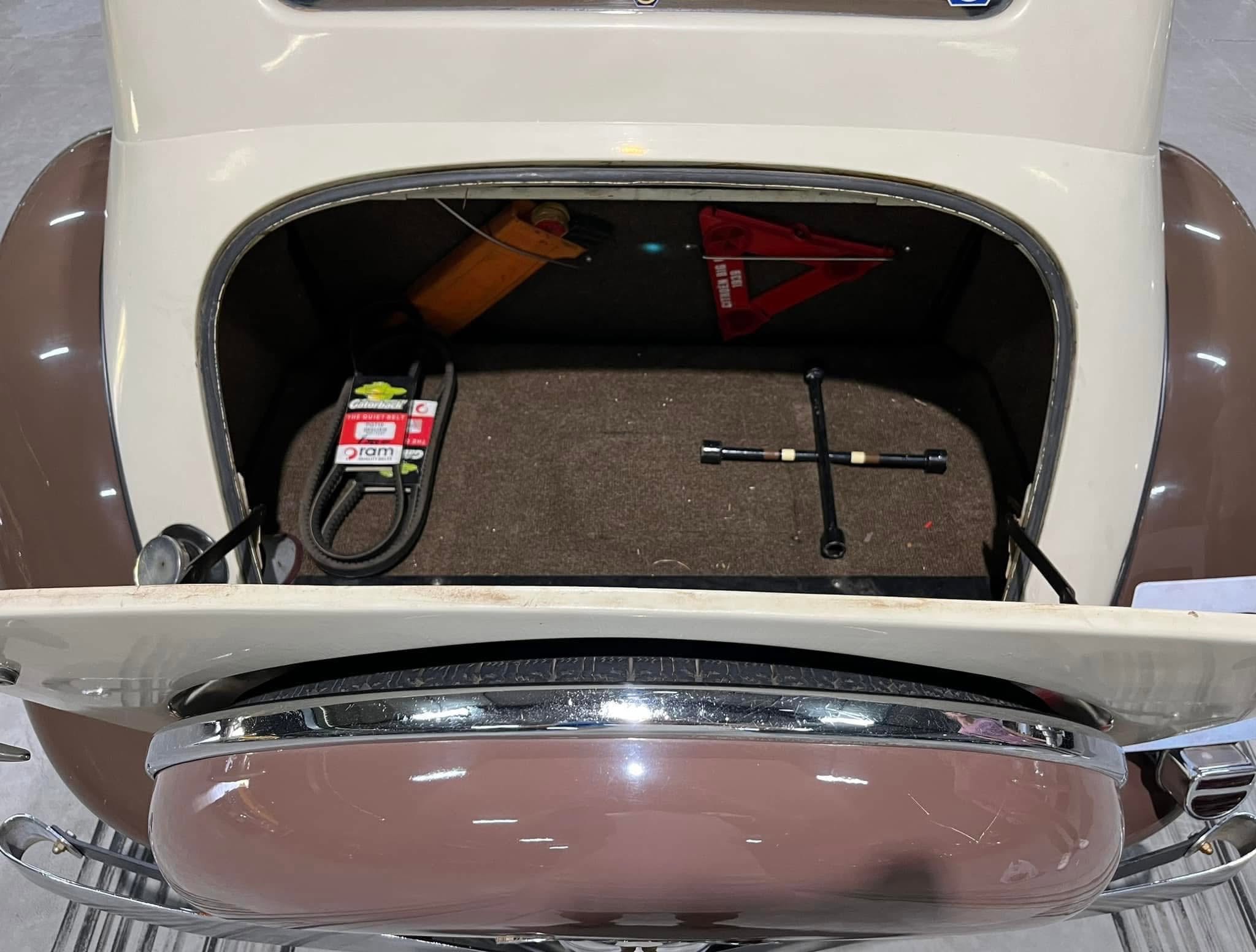 Open car trunk showing storage area with a battery, tools, and a red warning triangle inside a vintage-style vehicle trunk.