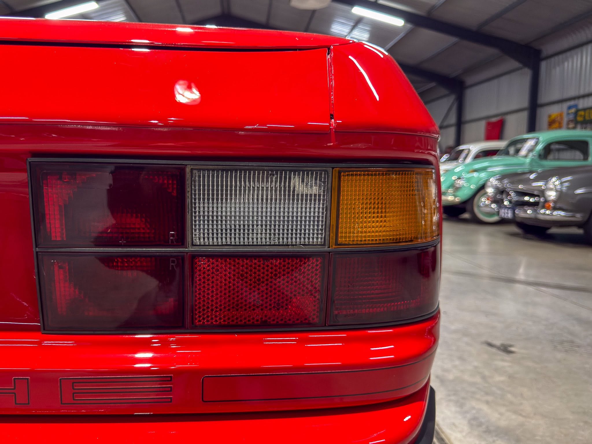 Close-up of the rear end of a red car, showing taillights and amber turn signal in a showroom with other vintage cars in the background.