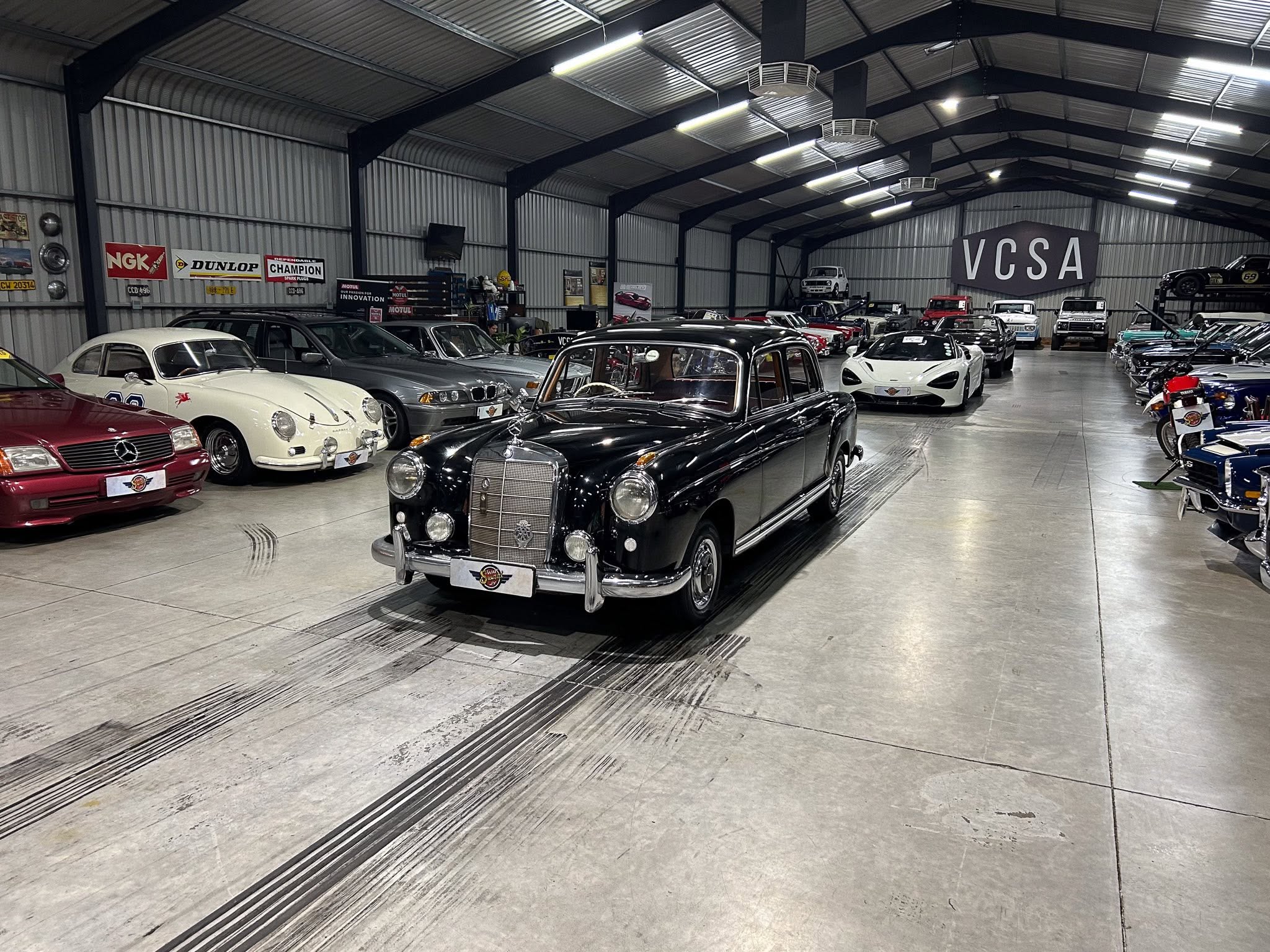Inside a large showroom filled with vintage and luxury cars parked in rows under a metal roof; signs along the wall indicate sponsors.