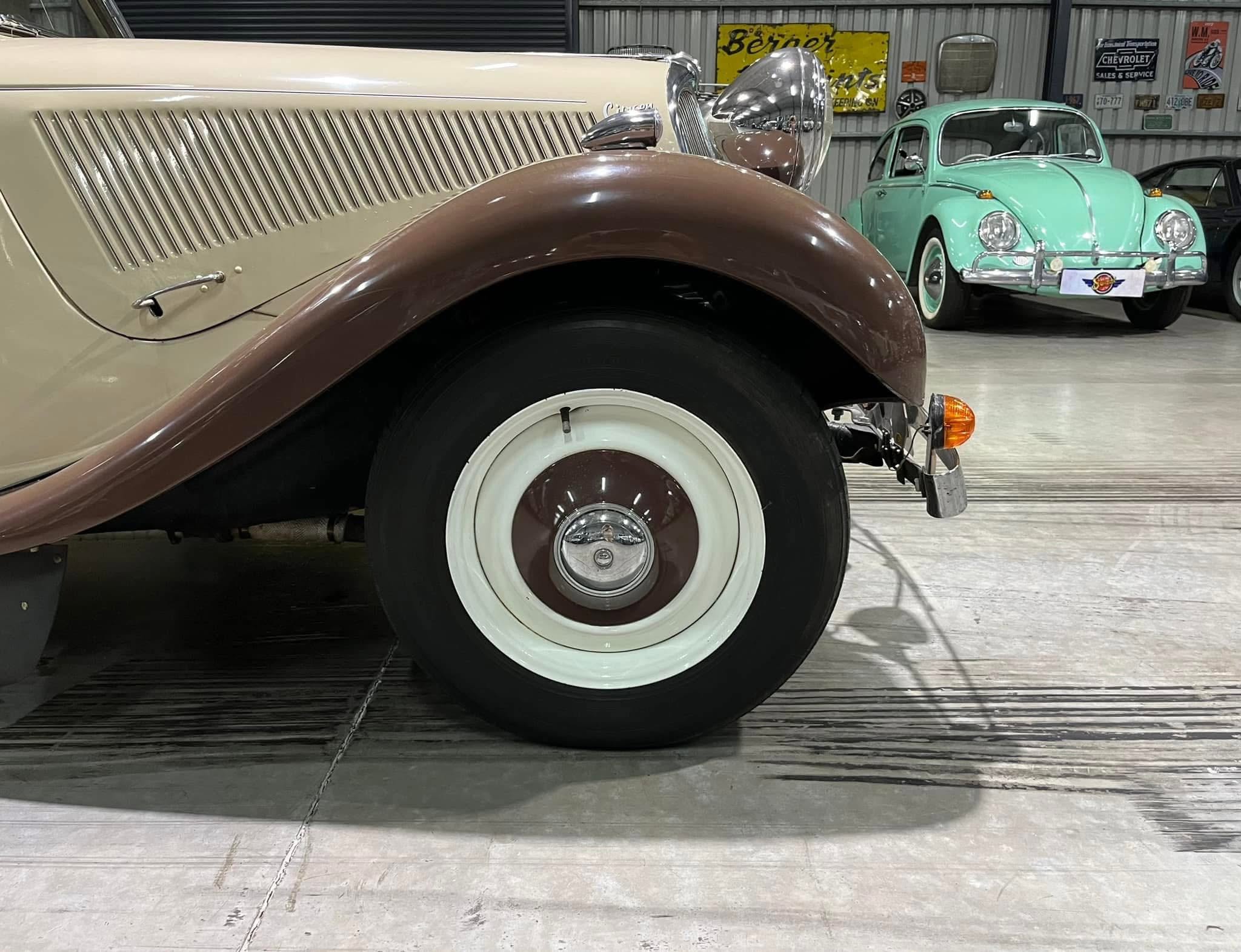 Close-up of a vintage beige car's front wheel and brown fender inside a showroom. In the background is a mint-green classic Volkswagen Beetle.