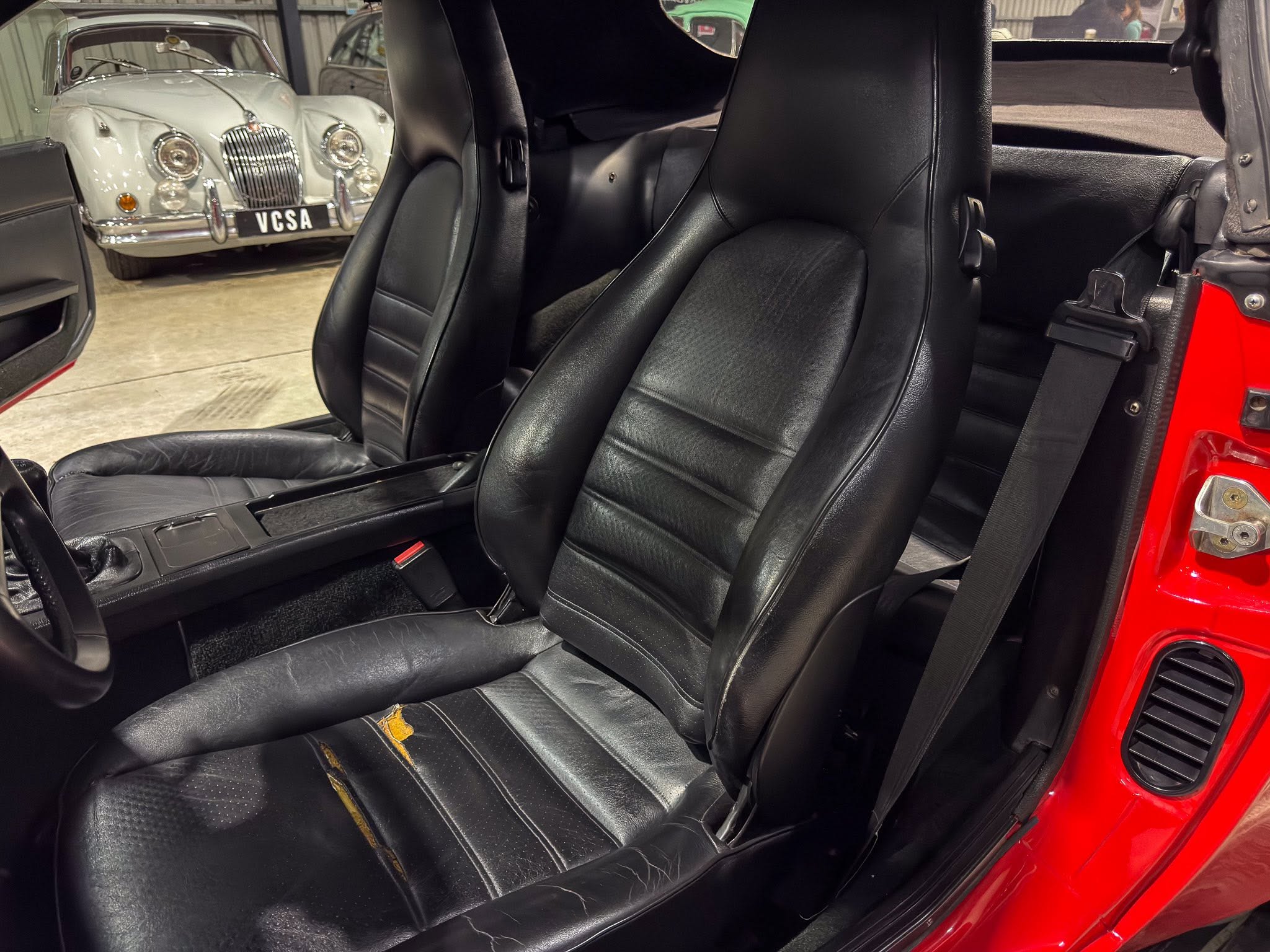 Black leather car seats inside a red vehicle, with a classic white car visible through the open door in the background.