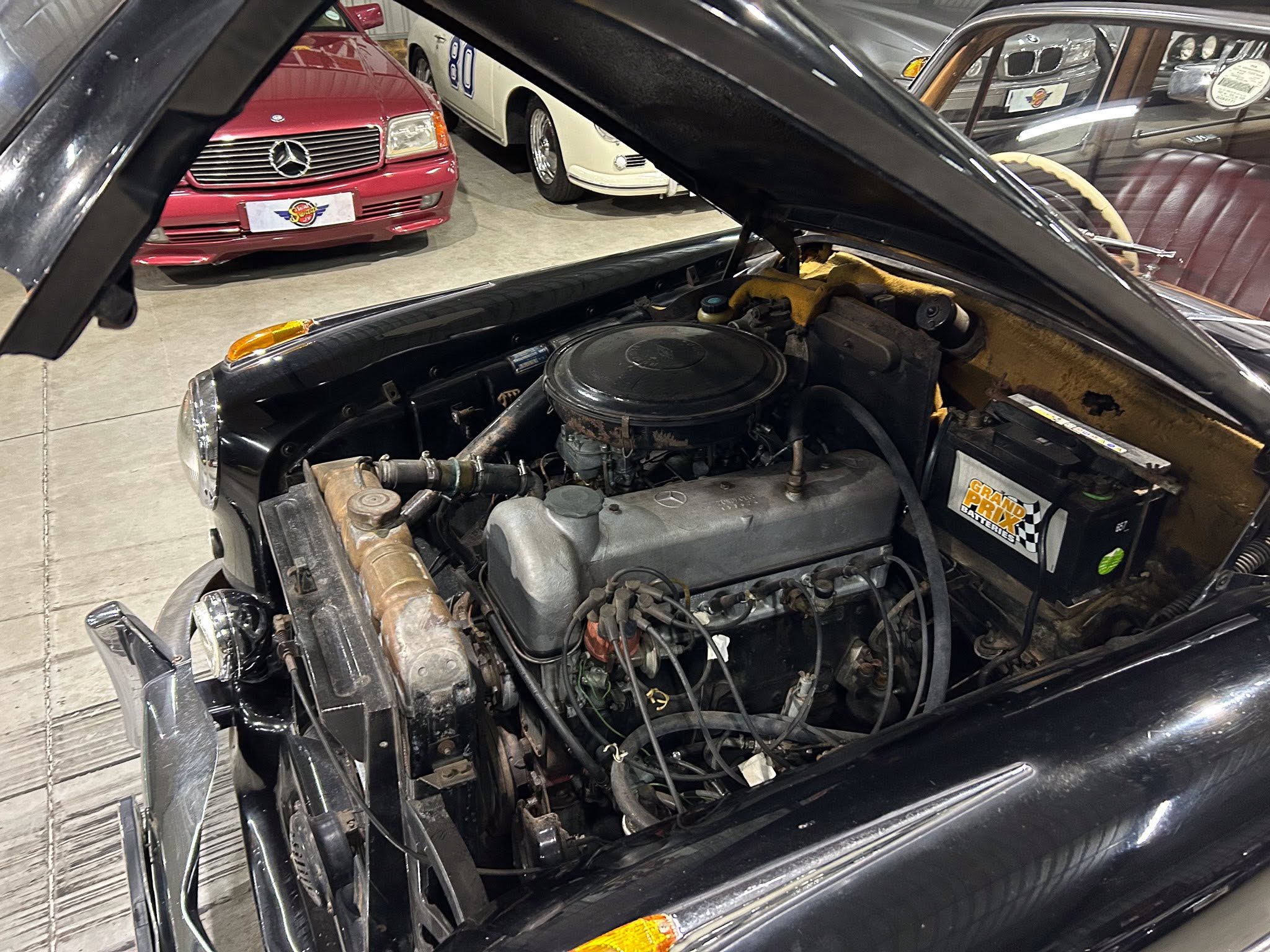 Open car hood exposing engine bay in a classic car showroom with another red Mercedes and white vehicles in the background