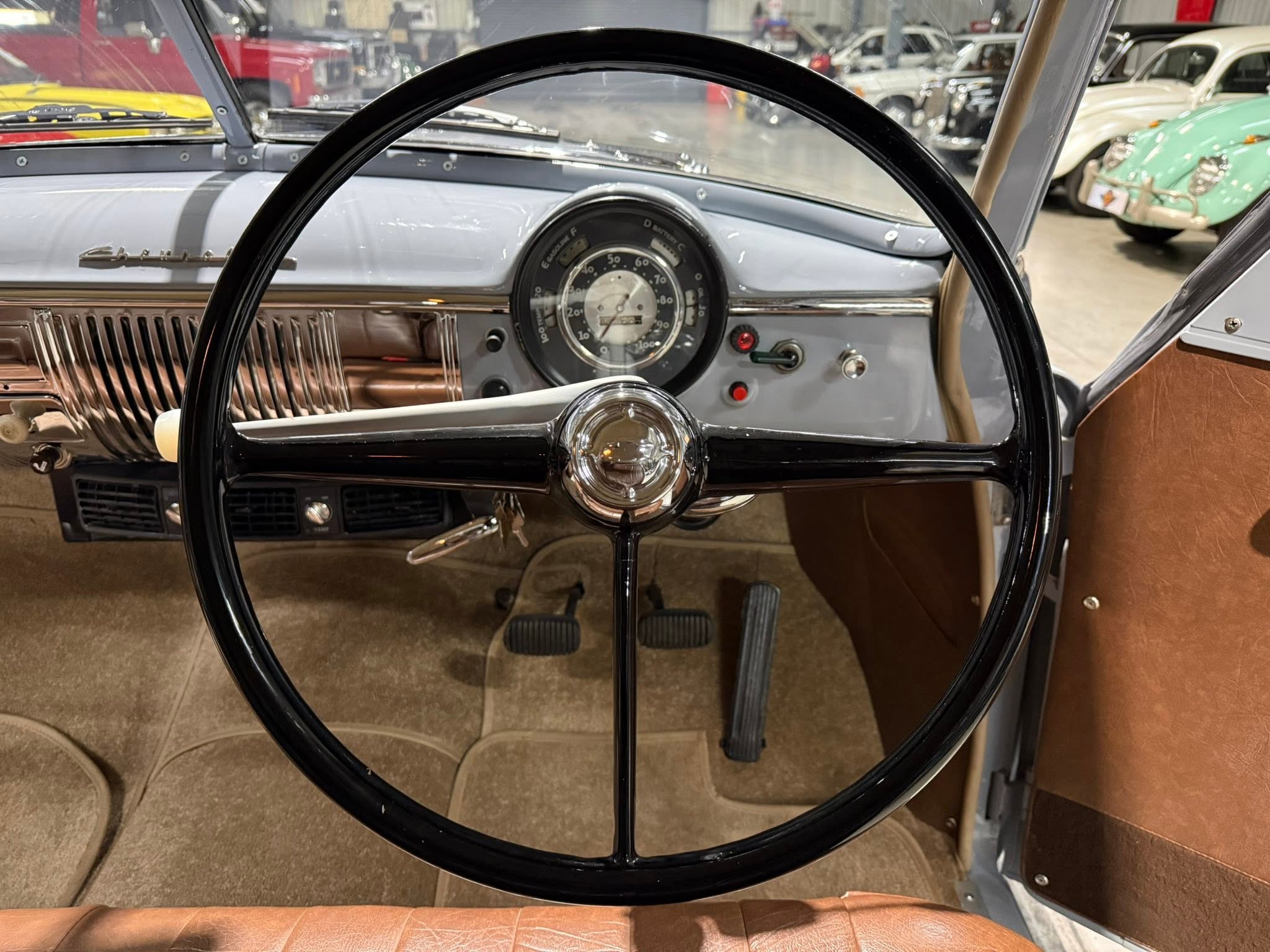 Classic car interior with a large circular instrument gauge centered behind a black steering wheel, brown seat upholstery visible to the right, showroom in background