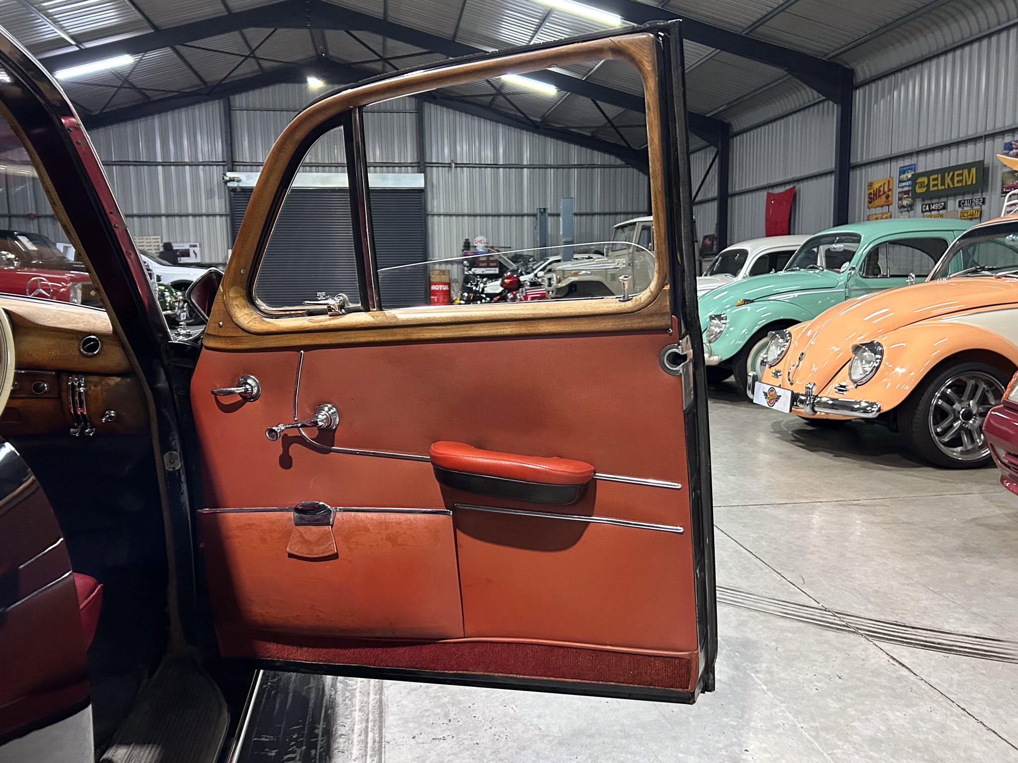 Open car door showing brown leather and wood trim inside a vintage car, with classic cars on display in a showroom behind.