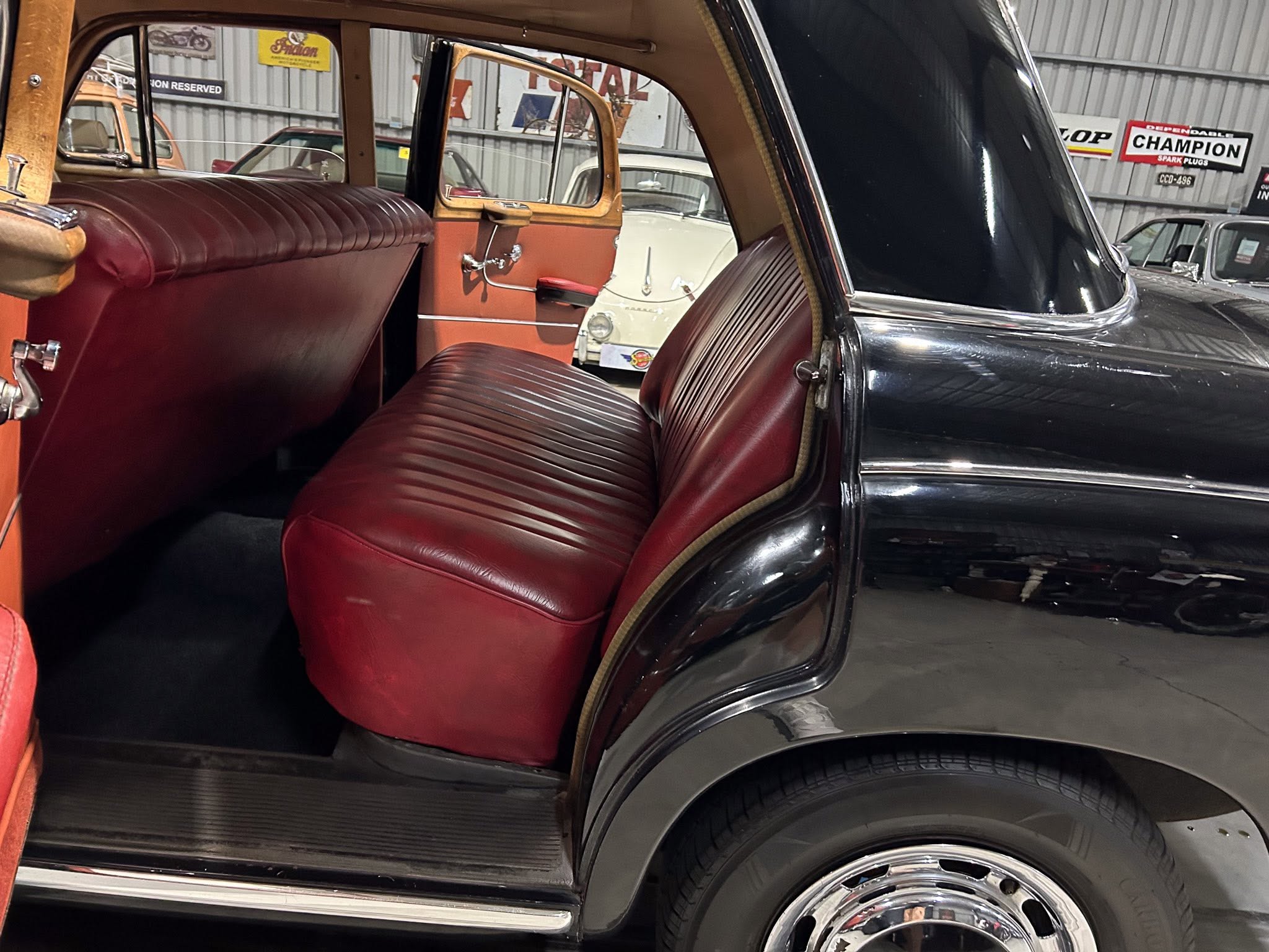 Interior of a classic car with red pleated leather seats and wood trim, open rear door, in a showroom with other vintage cars visible outside the window.
