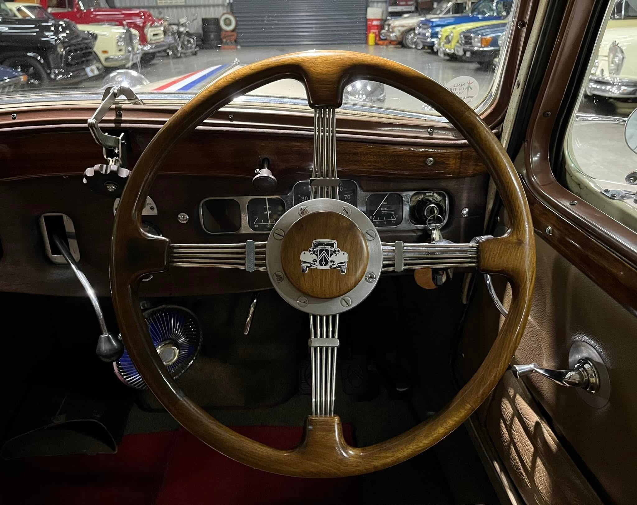 Vintage car interior with a wooden steering wheel, chrome spokes, and retro gauges; classic cars visible through the windshield in a showroom.