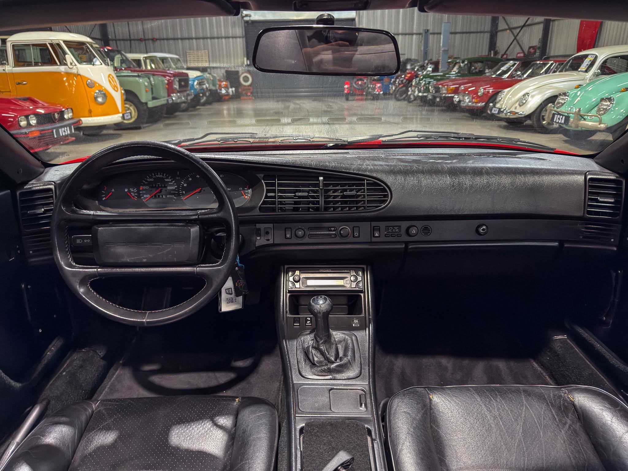 From the driver’s seat, the dashboard and gear shifter are visible while colorful vintage cars line up in a showroom viewed through the windshield.