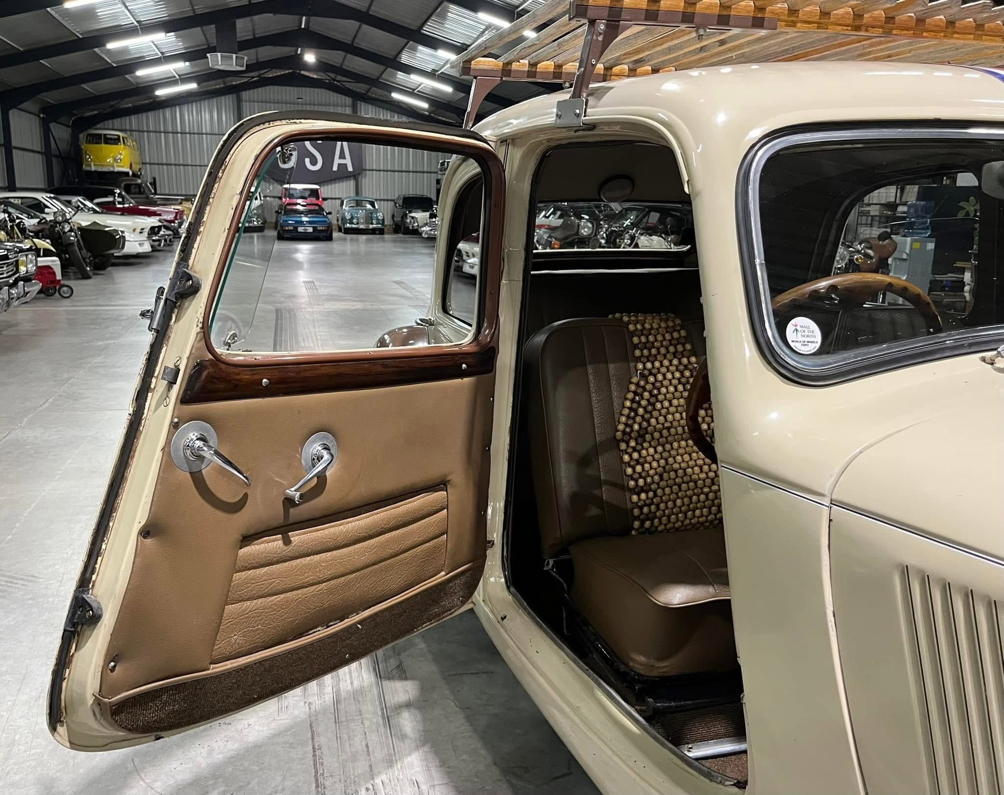 Open vintage car door showing tan interior and bead seat cover inside a showroom full of classic cars behind glass, with a wooden rack on top of the car.