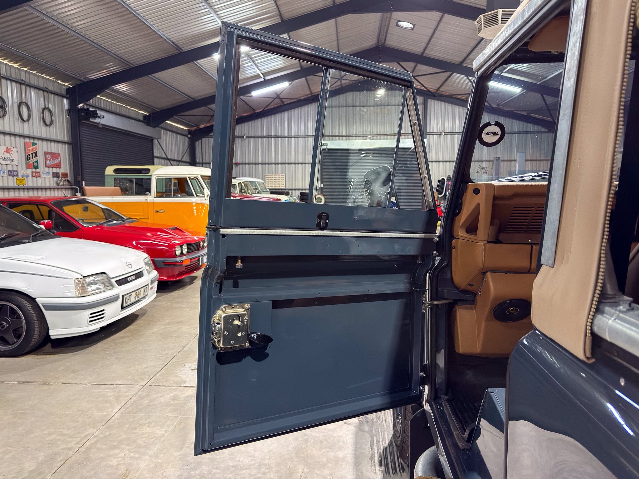 Open rear door of a dark blue SUV inside a vintage car showroom, with classic cars parked along the walls in a metal building.