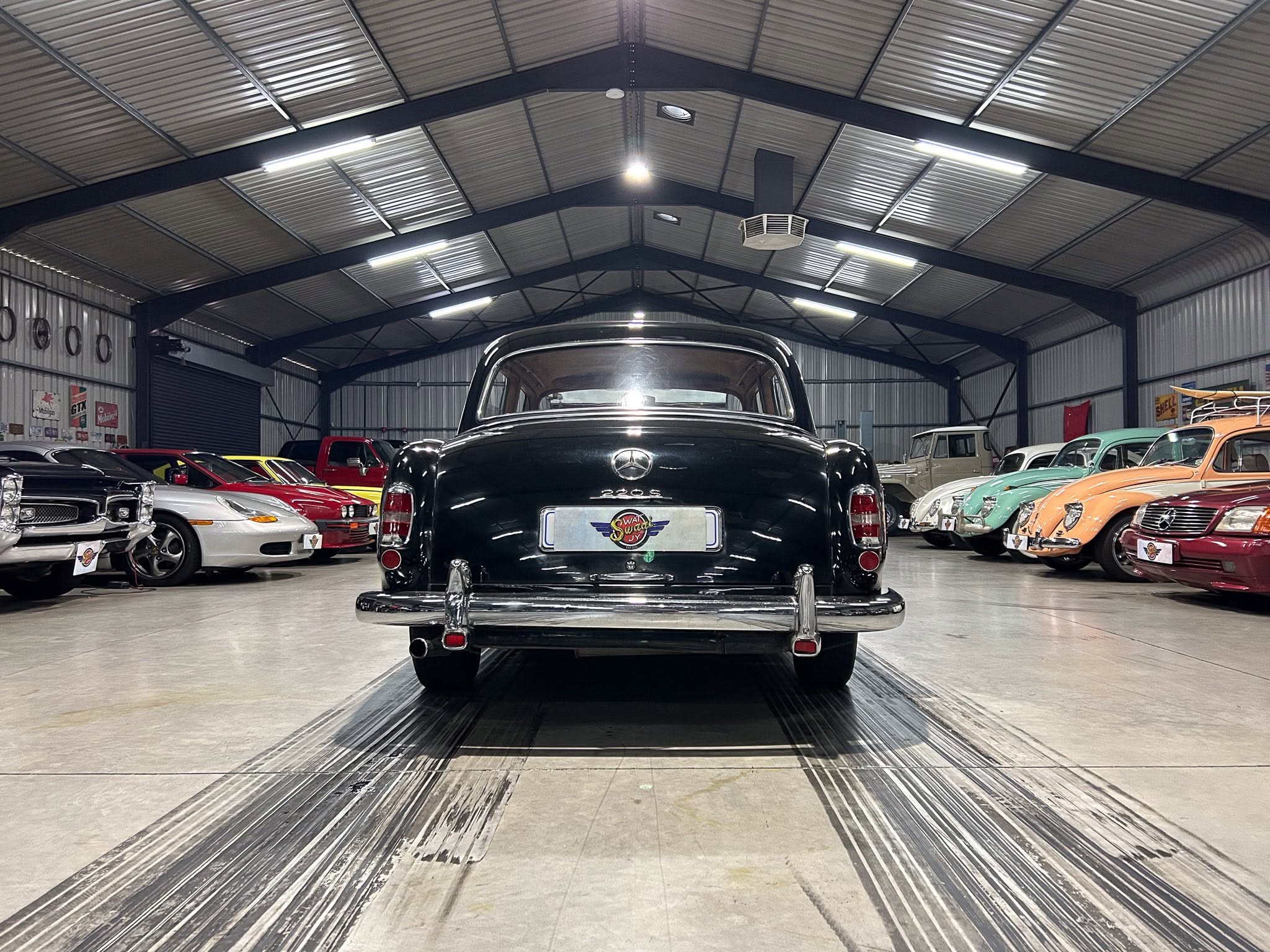 Rear view of a black vintage Mercedes-Benz 220S in a museum showroom with other classic cars on both sides.