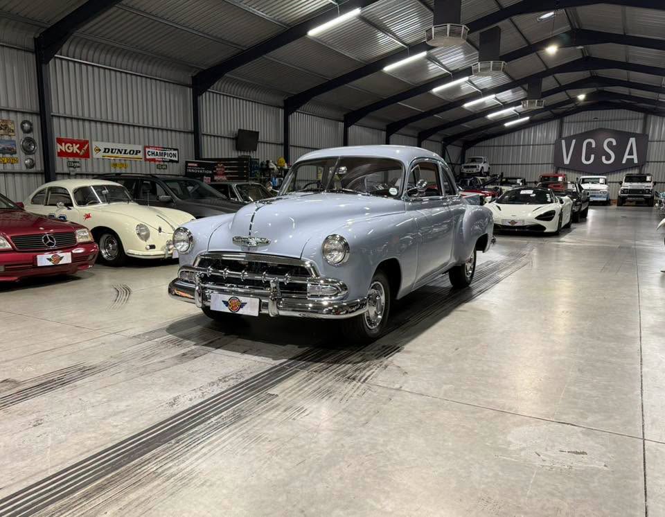 Blue 1950s-era vintage car on display in a large indoor showroom with other classic cars nearby.
