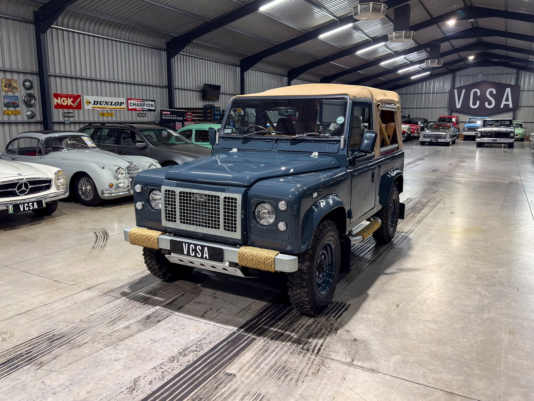 Indoor showroom of classic cars; blue SUV with beige soft top in foreground, others lined along walls in the background.
