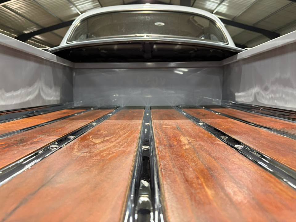 Low-angle view of a vintage pickup truck bed with wooden floor planks and metal rails, leading to the cab inside a warehouse.
