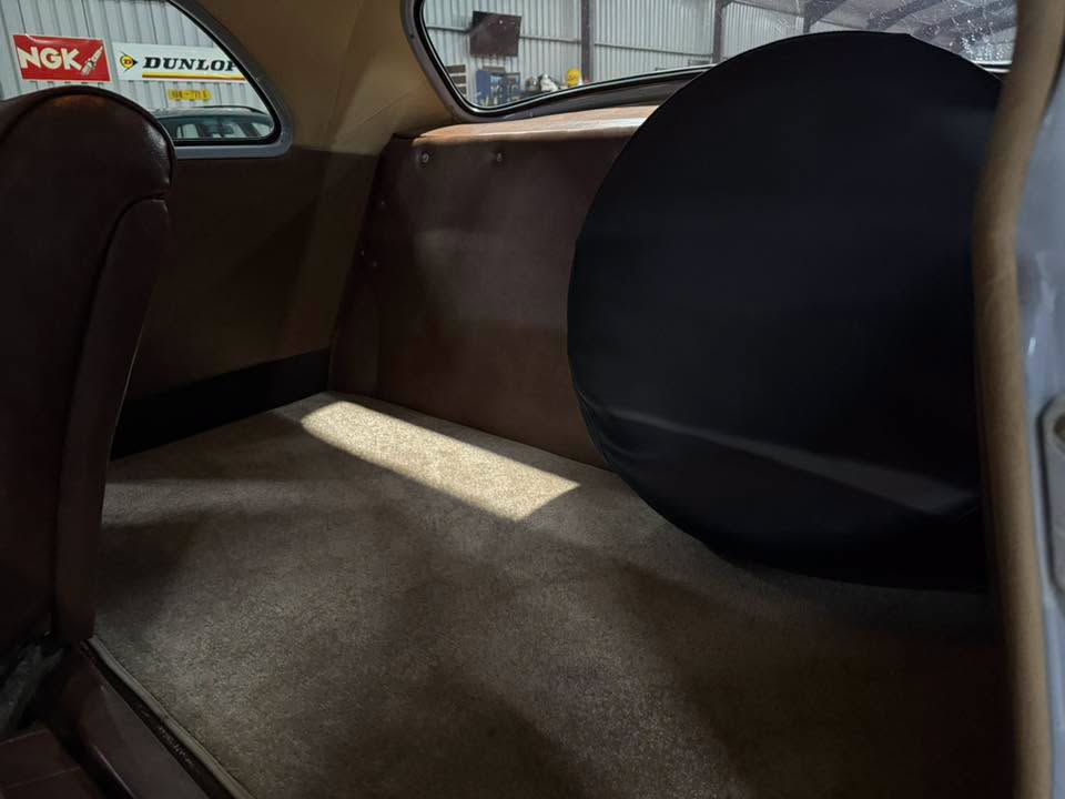 Interior of a vintage car showing the rear cargo area with a round black spare tire cover on the right and brown panels; sunlight on the carpeted floor in a garage setting.